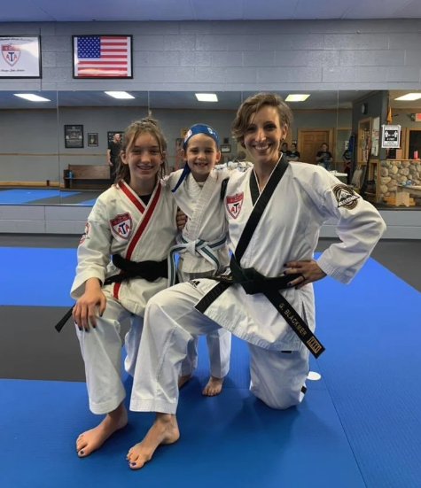 Three smiling karate students wearing white uniforms and competition medals kneel together on a blue training mat in a dojo, with an American flag visible on the wall.