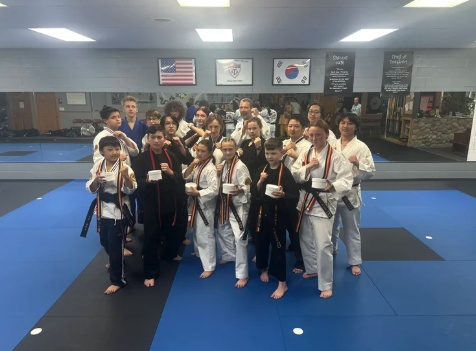 A large group of martial artists wearing white and black gis, many with medals, pose together on a blue mat inside a dojo with American and Korean flags on the walls.