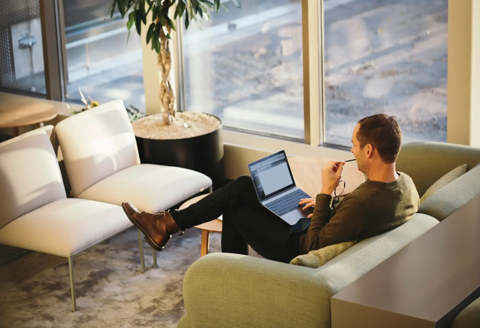Man working on a laptop in a lounge chair by tall windows