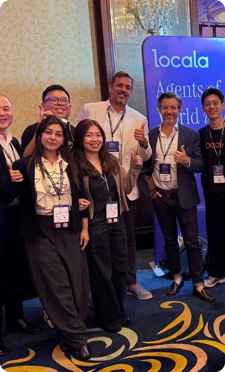 Group of seven diverse professionals standing indoors on a patterned carpet in front of a lit blue display sign that reads 'locala Agents of World'. They are smiling and giving thumbs-up gestures, wearing conference badges and business casual attire.