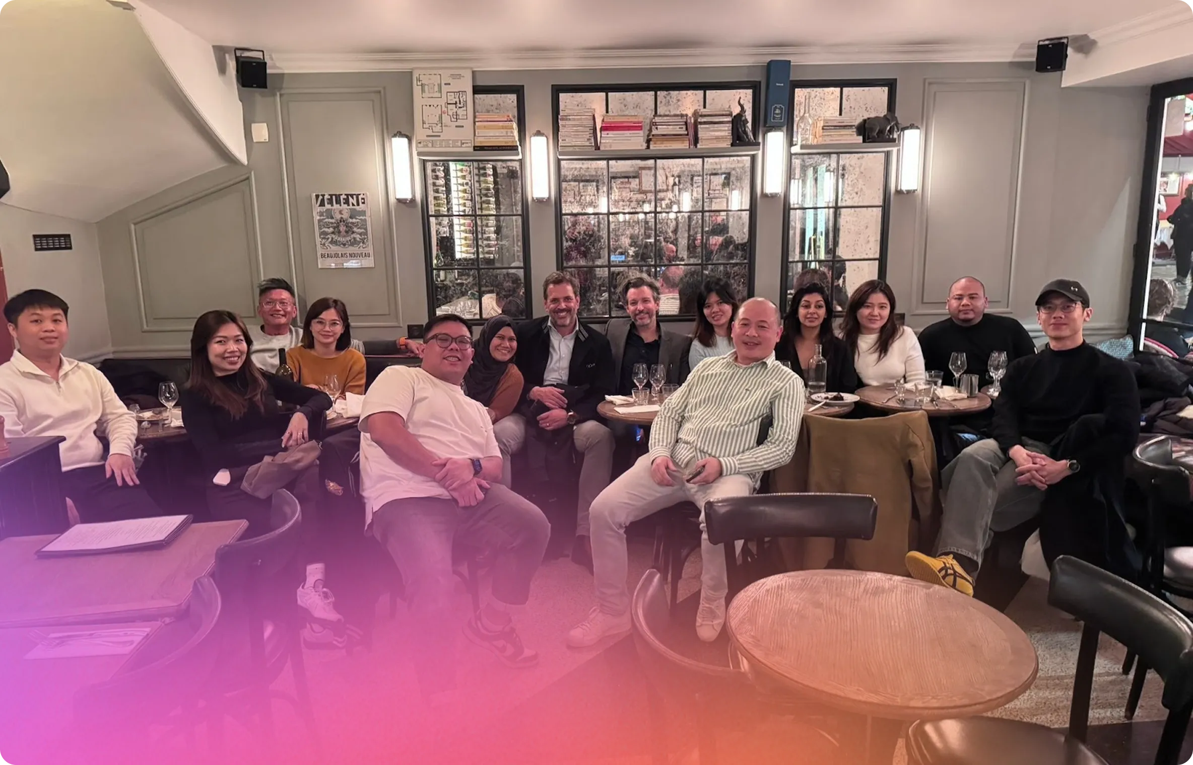 Group of thirteen people sitting closely together in a cozy restaurant with tables, chairs, and a mirror behind them reflecting the room.