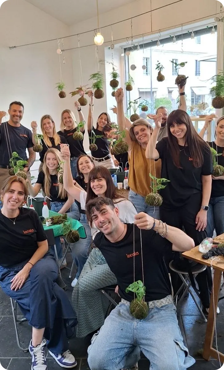 Group of smiling people indoors holding small hanging plants with roots wrapped in moss, appearing to be in a workshop or social gathering.