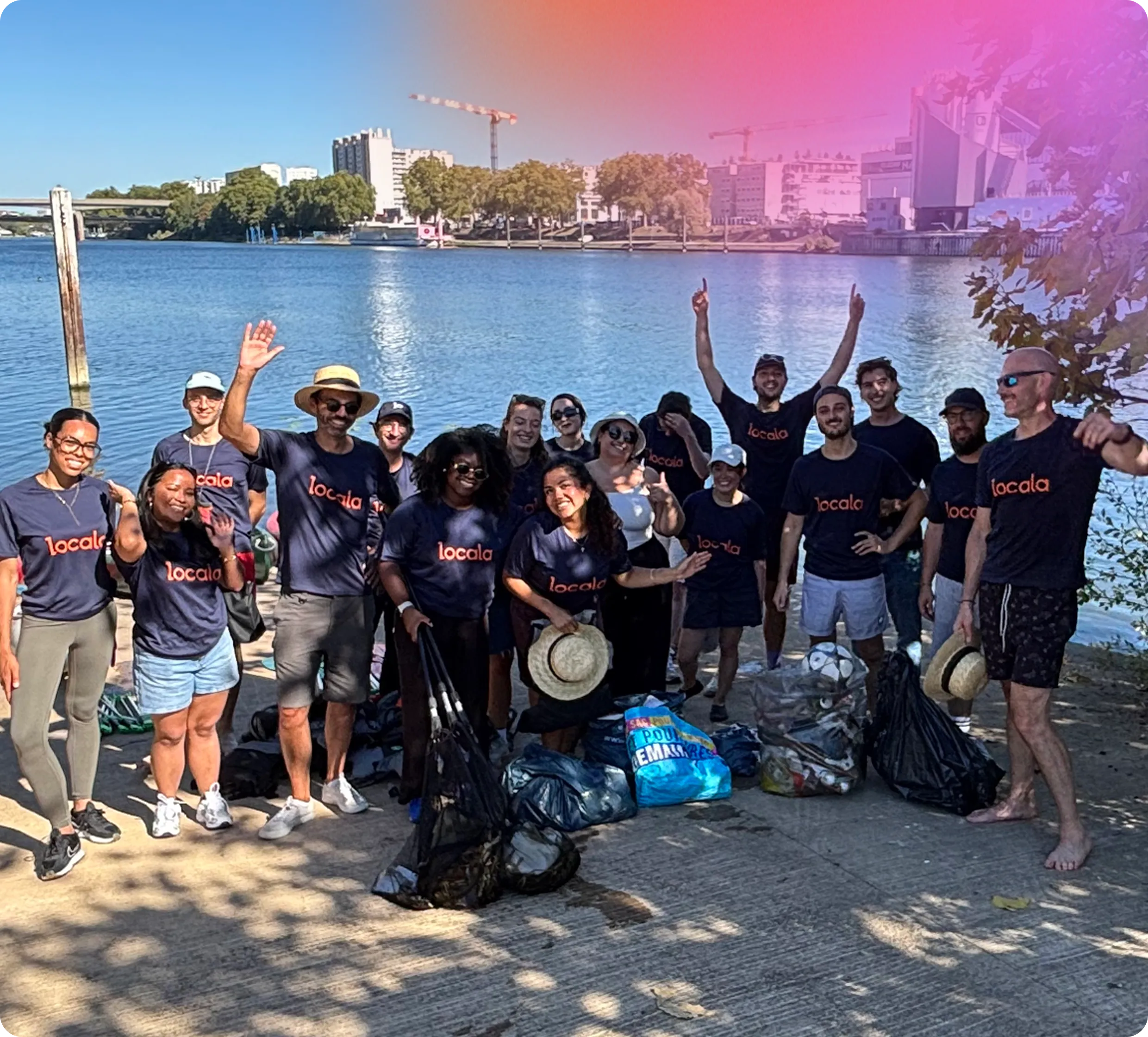 Group of diverse people wearing matching navy blue t-shirts with the word 'locala' printed on them, posing by a waterfront with trash bags filled with collected litter, some people smiling and waving, trees and buildings visible across the water.