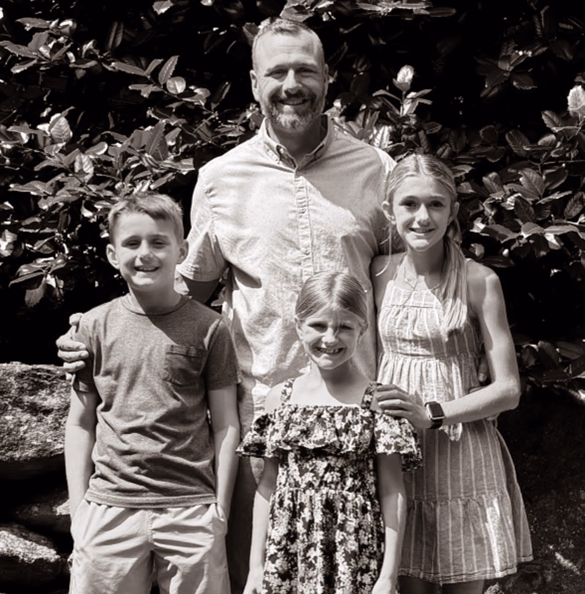 Smiling man stands with two girls and a boy outdoors in front of leafy bushes.