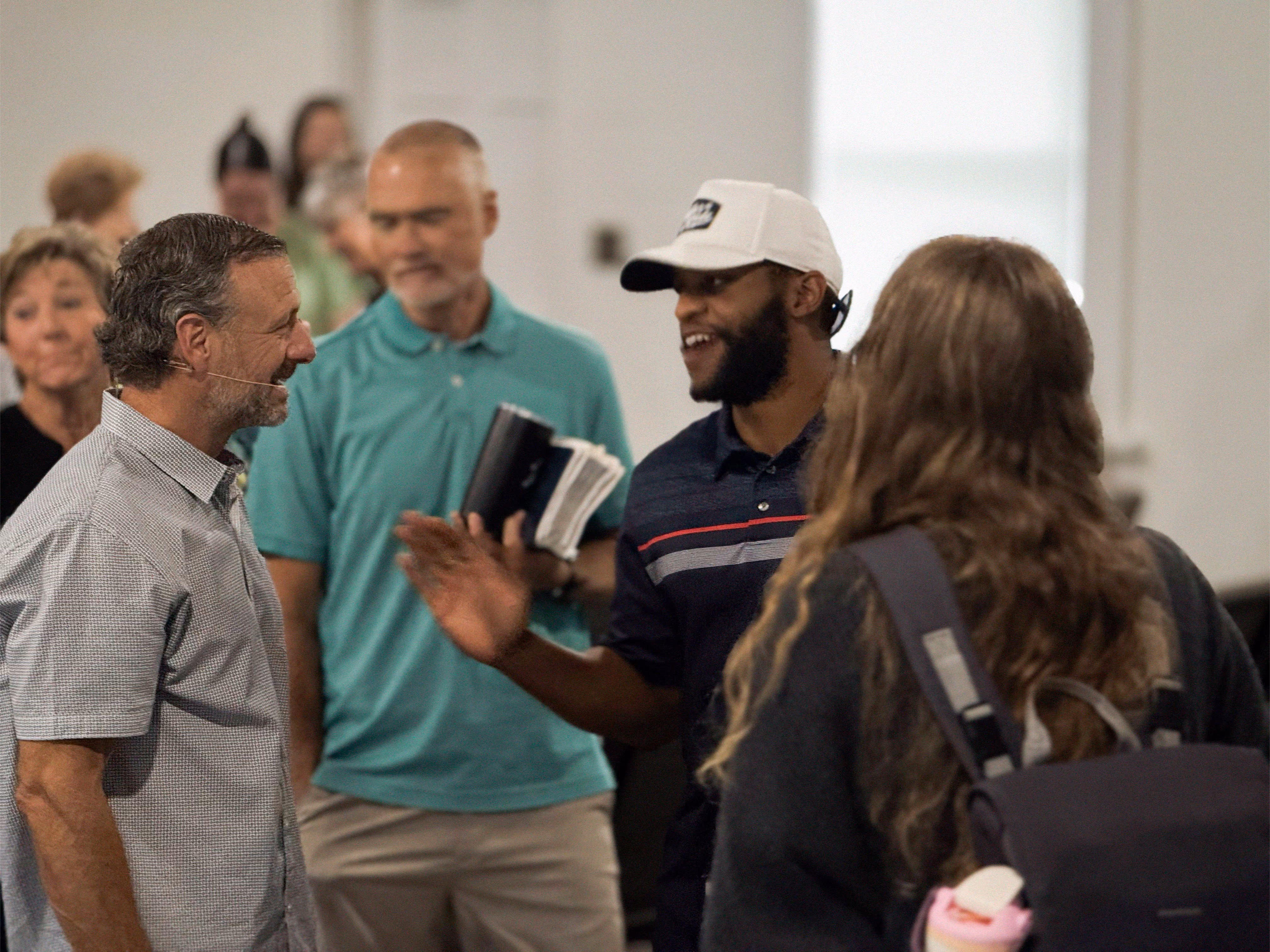 A group of people engaged in conversation inside a room, one man holding books and another wearing a white cap.