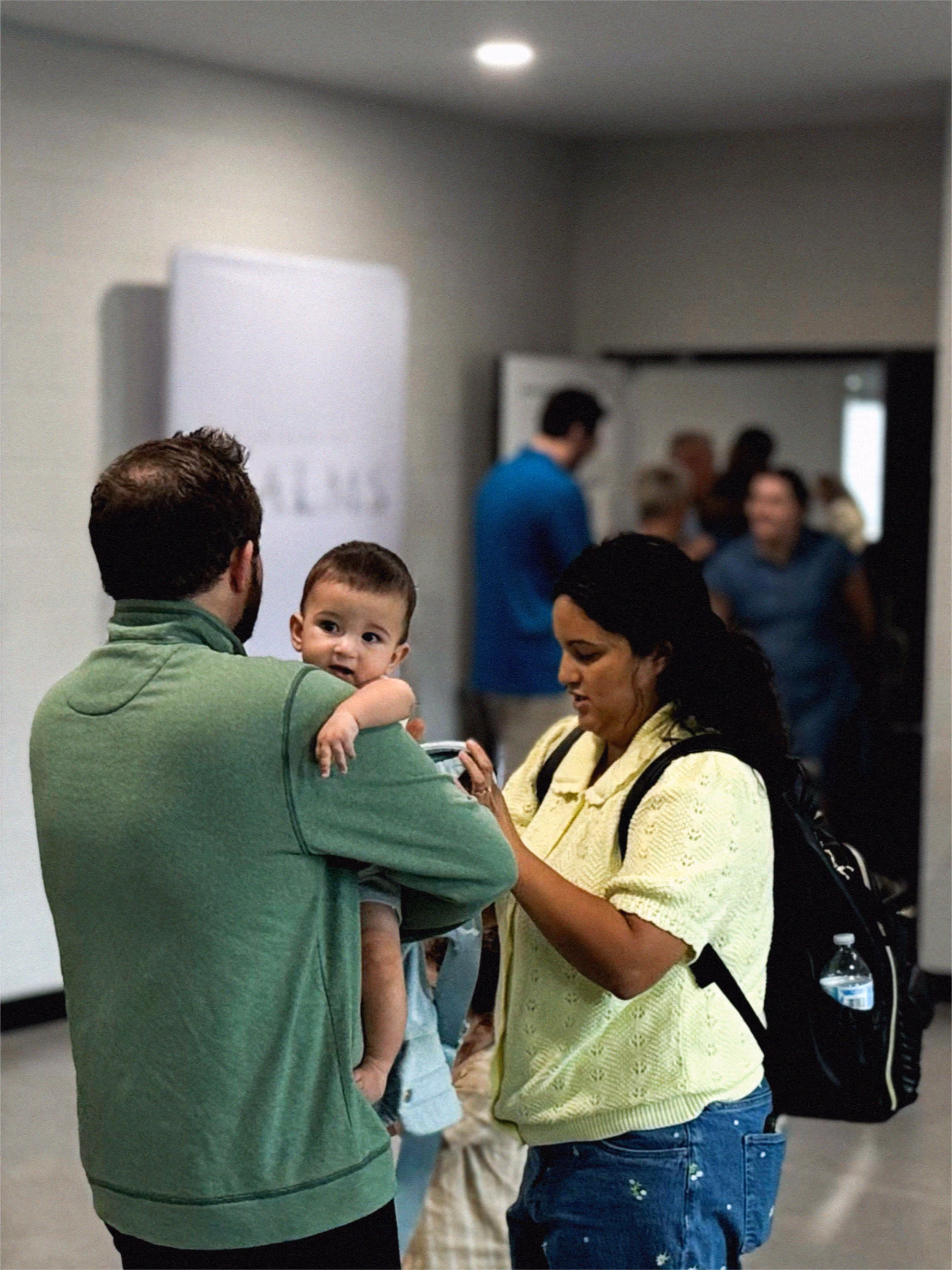 Man in green jacket holding baby while woman in yellow adjusts baby carrier indoors with people in background.
