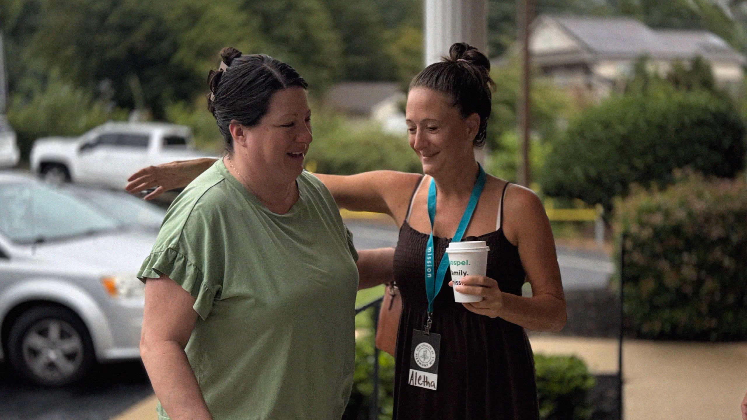 Two women smiling and embracing outdoors, one holding a coffee cup and wearing a name badge.