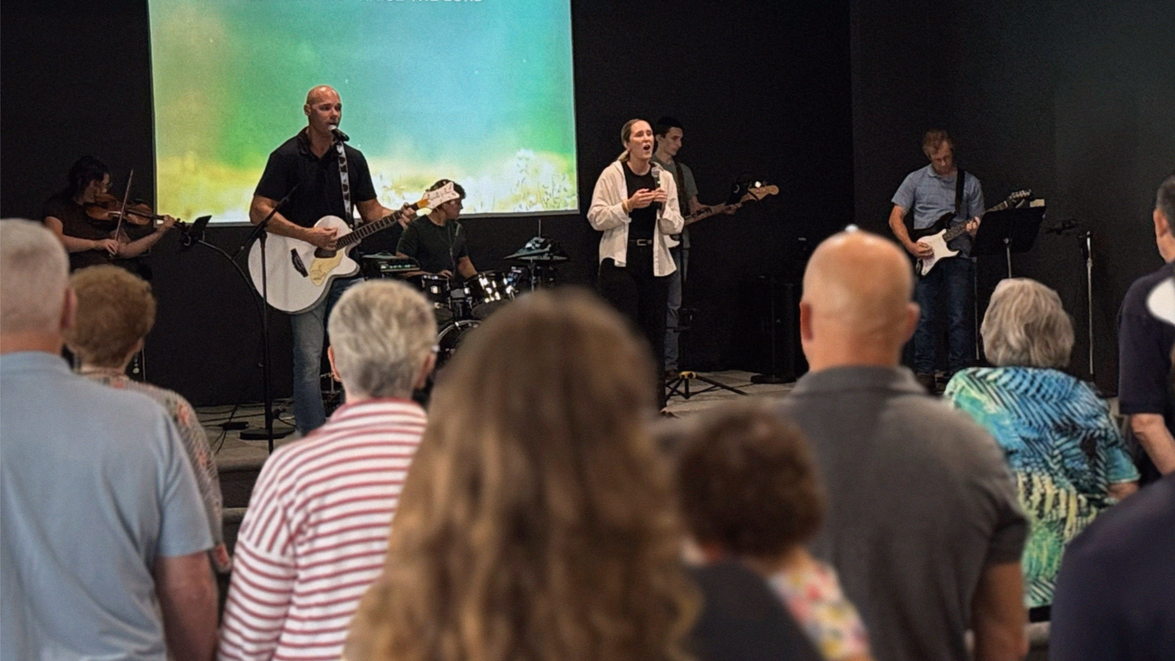 A band with guitar, violin, drums, and vocals performing in front of a seated audience.