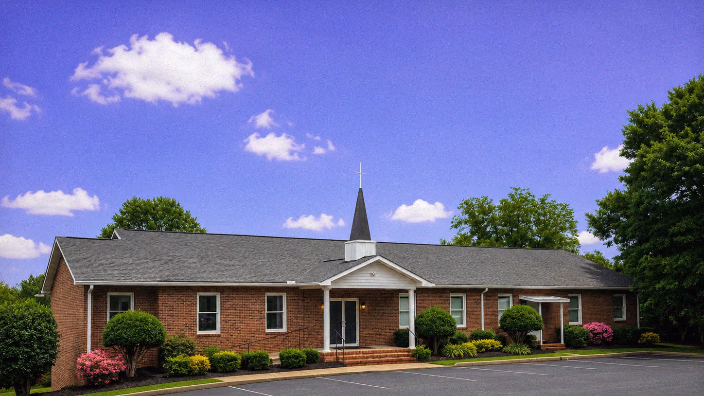 Brick church building with a steeple, surrounded by trees and bushes under a blue sky with clouds.