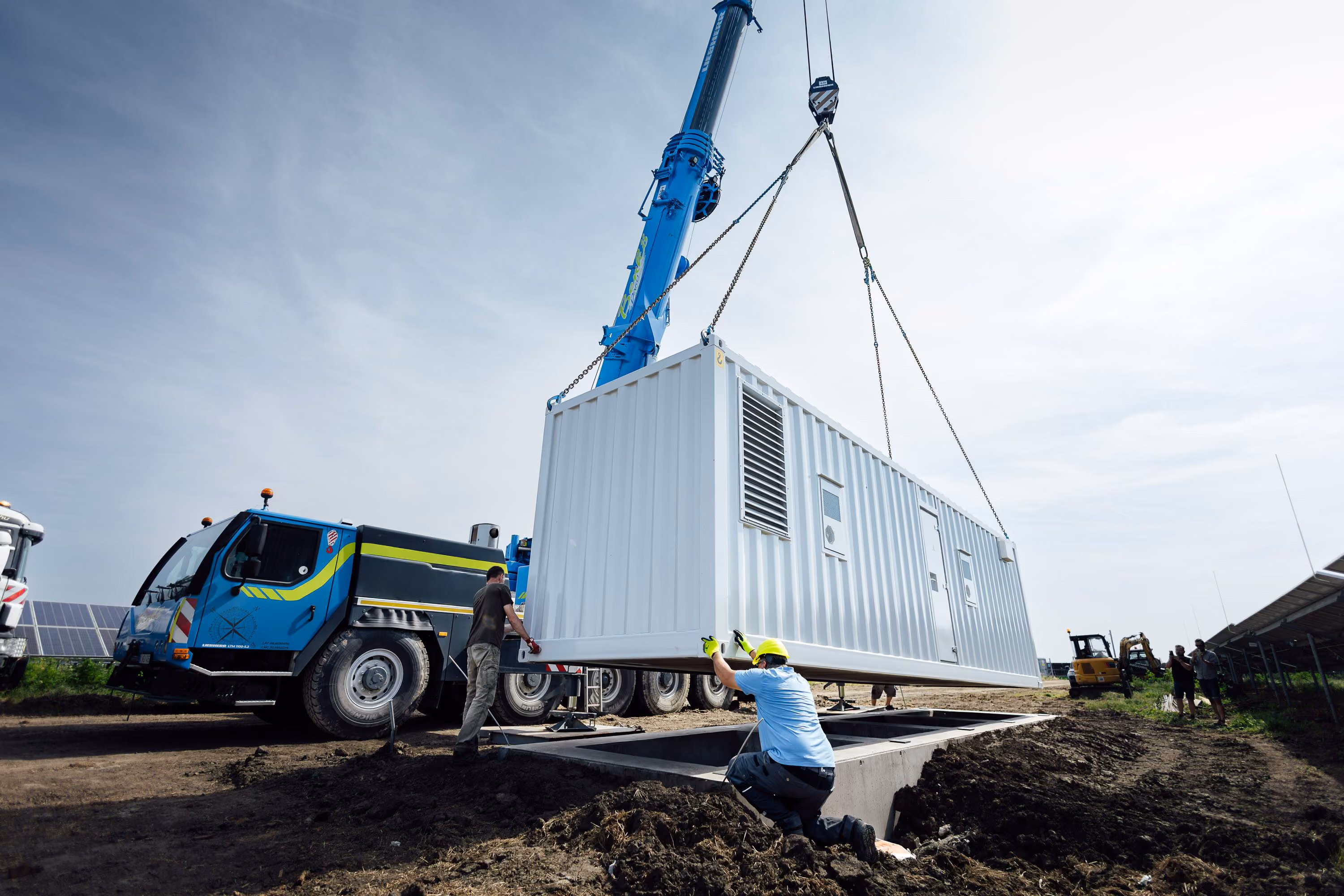 Jeřáb osazuje kontejnerové bateriové úložiště na betonový základ u fotovoltaické elektrárny.