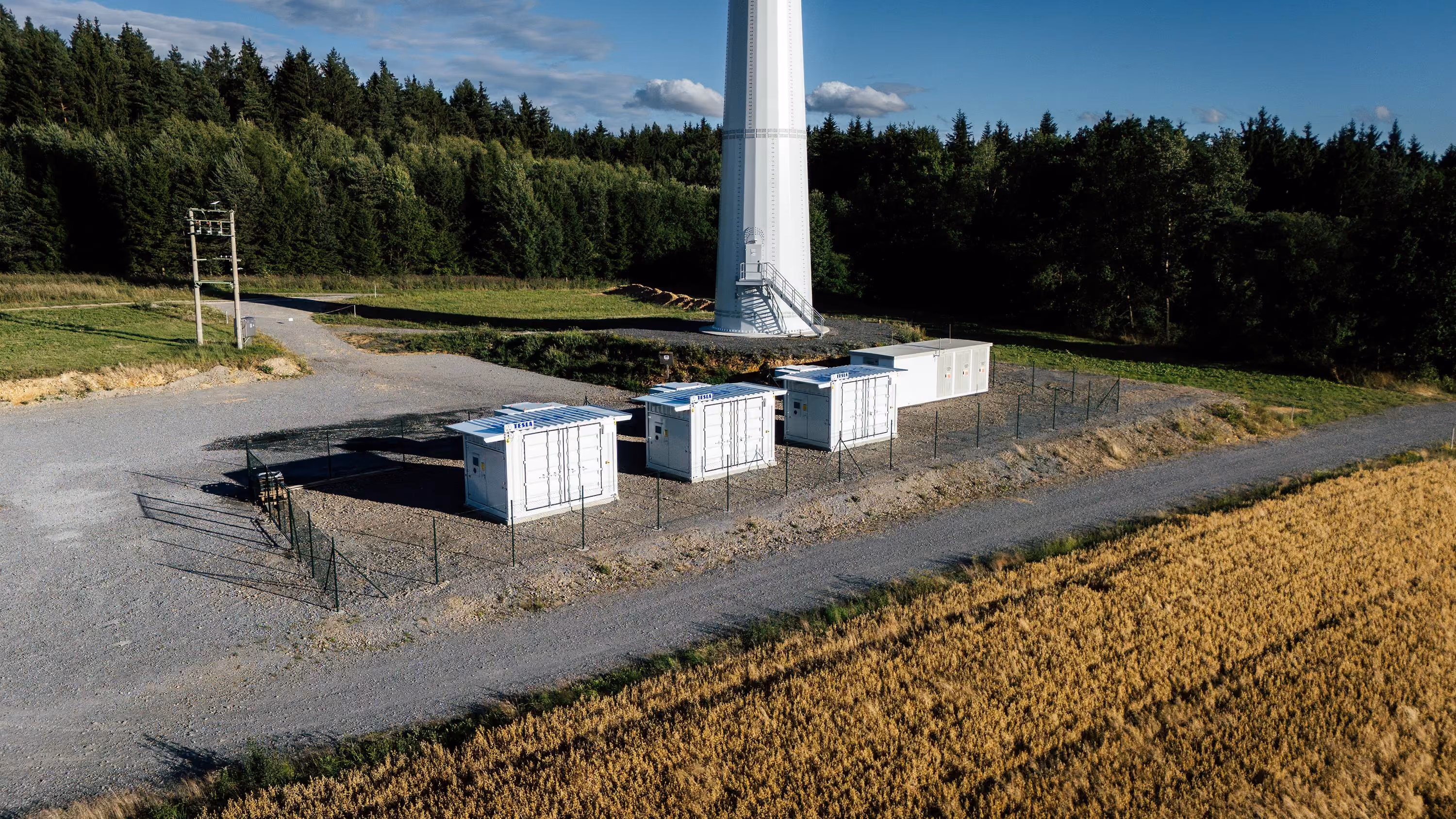 Letecký pohled na základnu větrné elektrárny se třemi bílými řídicími kontejnery obehnanými plotem, okolo jsou pole a les pod modrou oblohou s mraky.