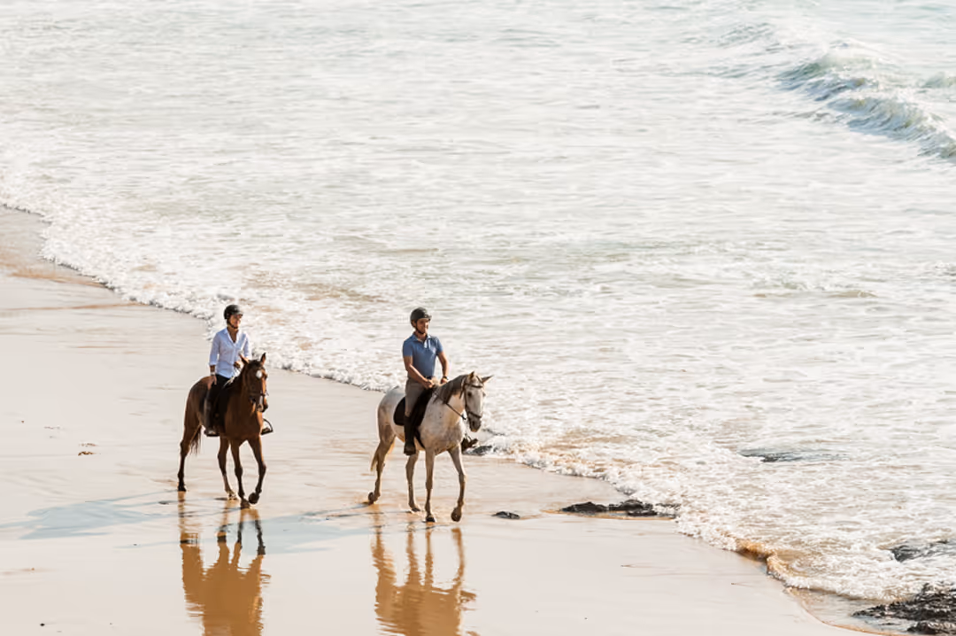 Two people wearing helmets enjoying a slow leisure horse ride along the scenic beach coastline.