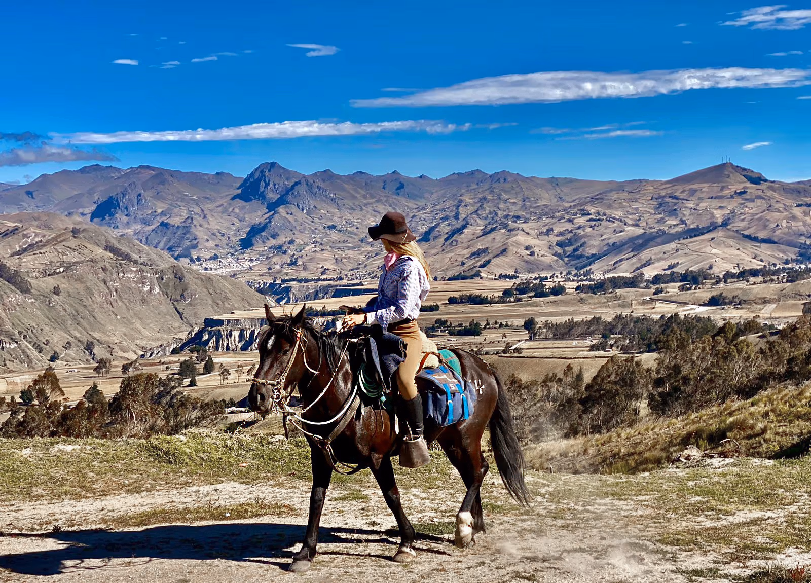Une cavalière aux longs cheveux blonds portant un chapeau de soleil regarde vers l’horizon et les paysages des Andes.