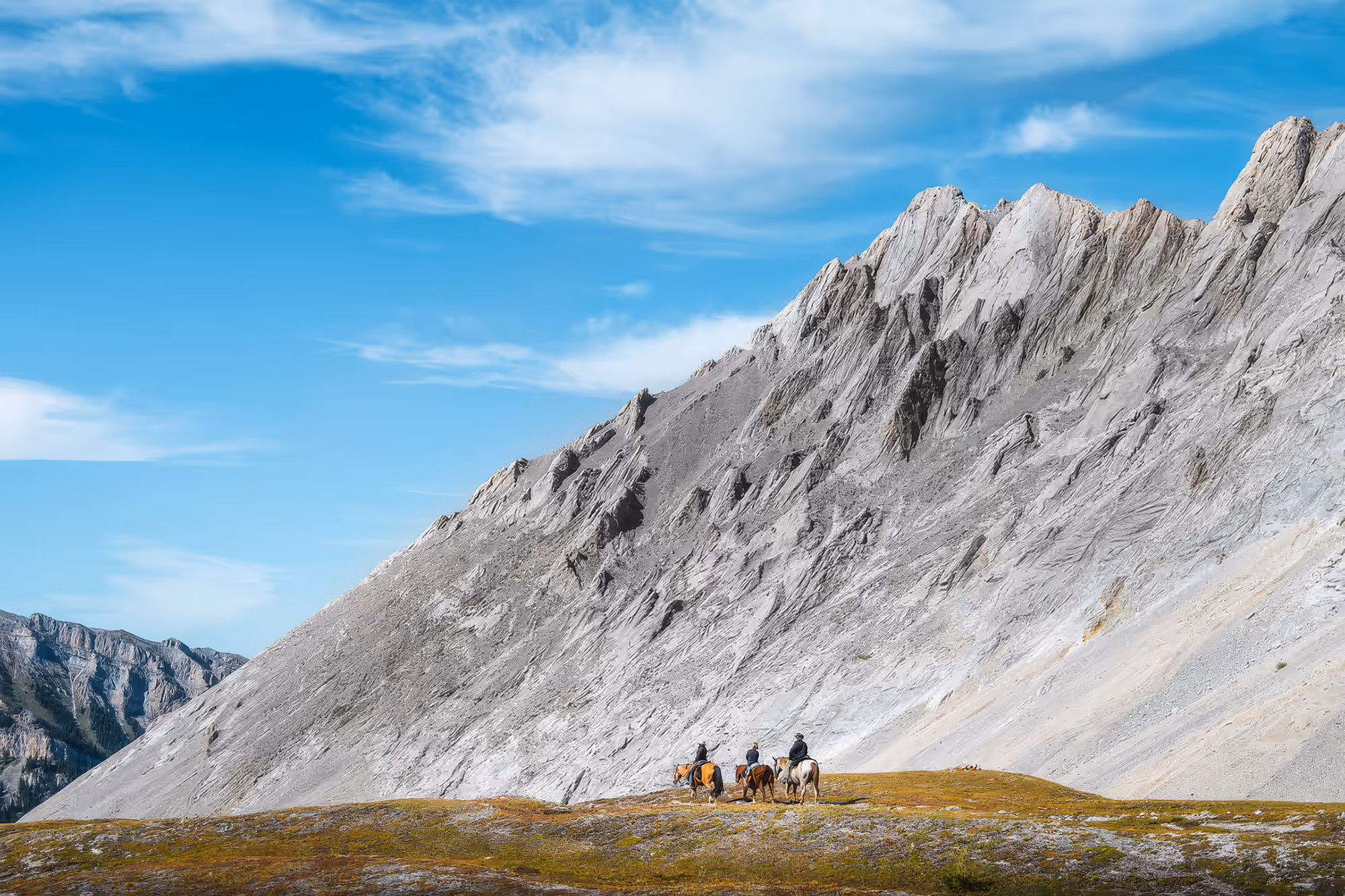 Vue panoramique d’un groupe de cavaliers traversant une vaste prairie verdoyante au pied d’une montagne rocheuse dénudée.