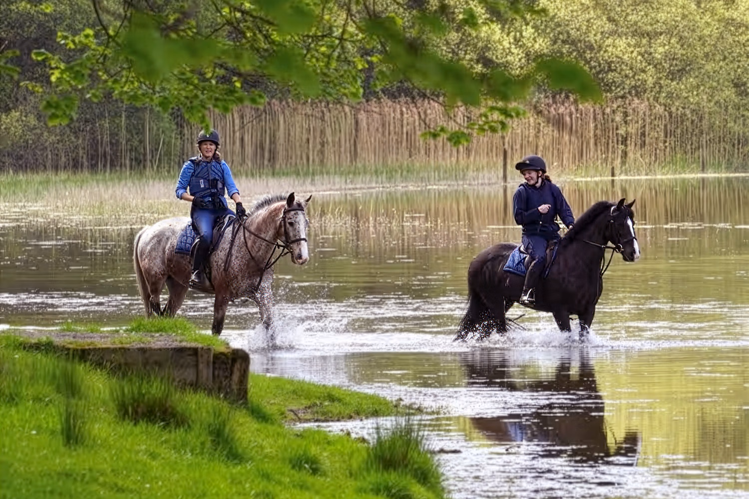Deux cavaliers traversant calmement les eaux peu profondes au bord d’un lac pittoresque.