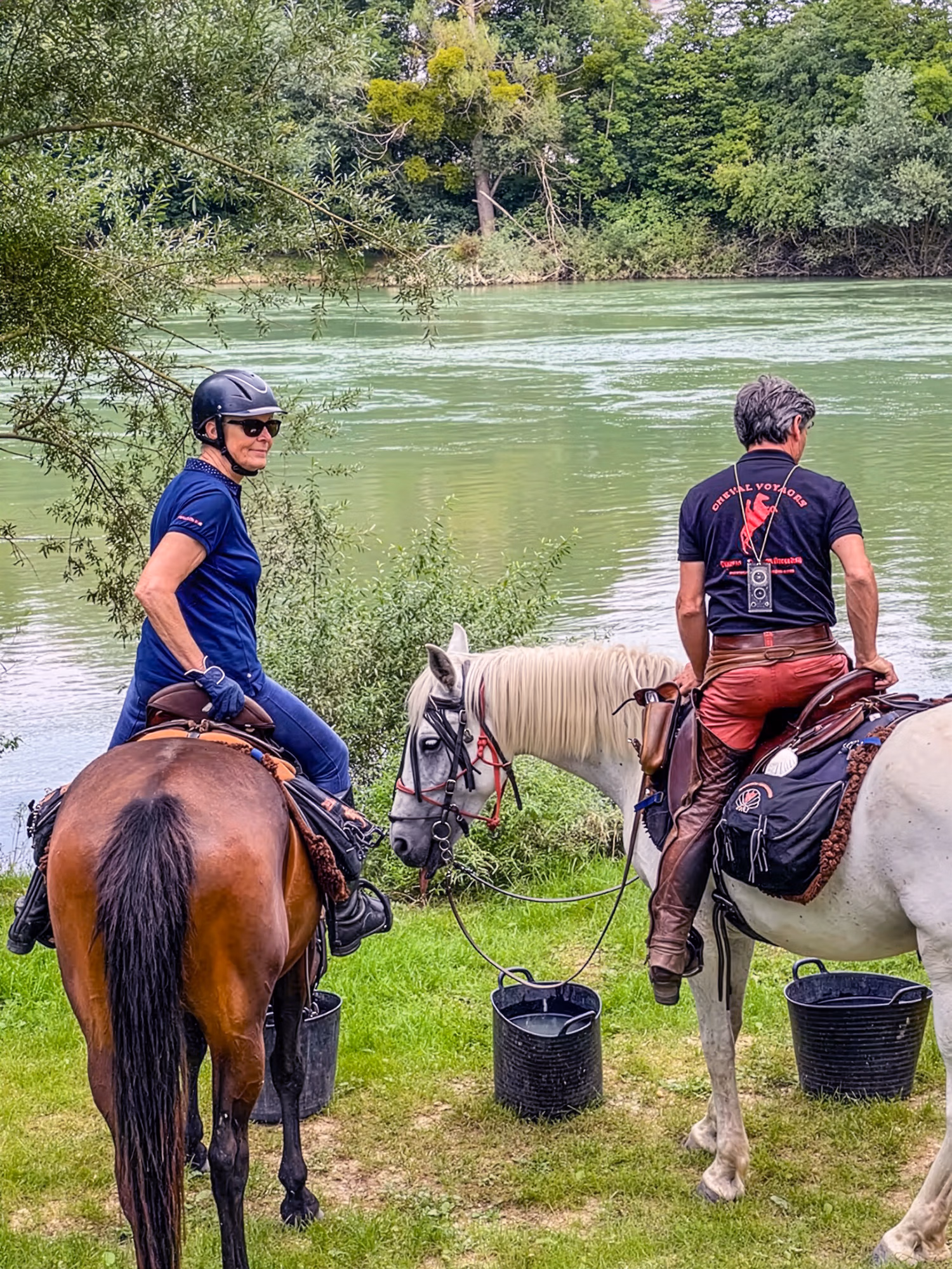 Deux cavaliers sur leurs chevaux qui prennent une pause pour se désaltérer dans des seaux au bord d'une riviere lors d'une randonnée équestre.