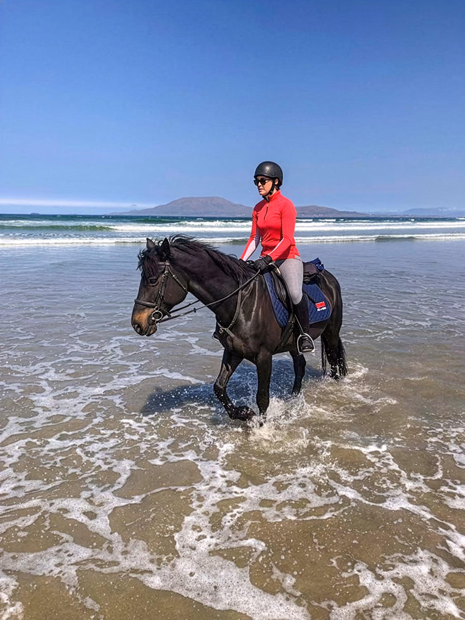 Une cavalière en haut rouge et portant un casque traverse l’eau avec son cheval.