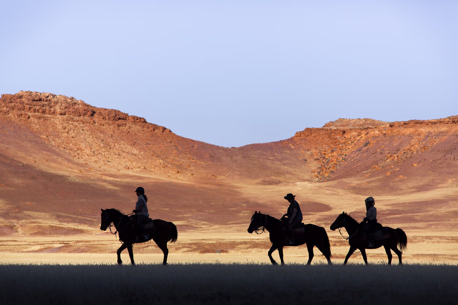 Silhouette de trois cavaliers avançant lentement devant un imposant paysage montagneux.
