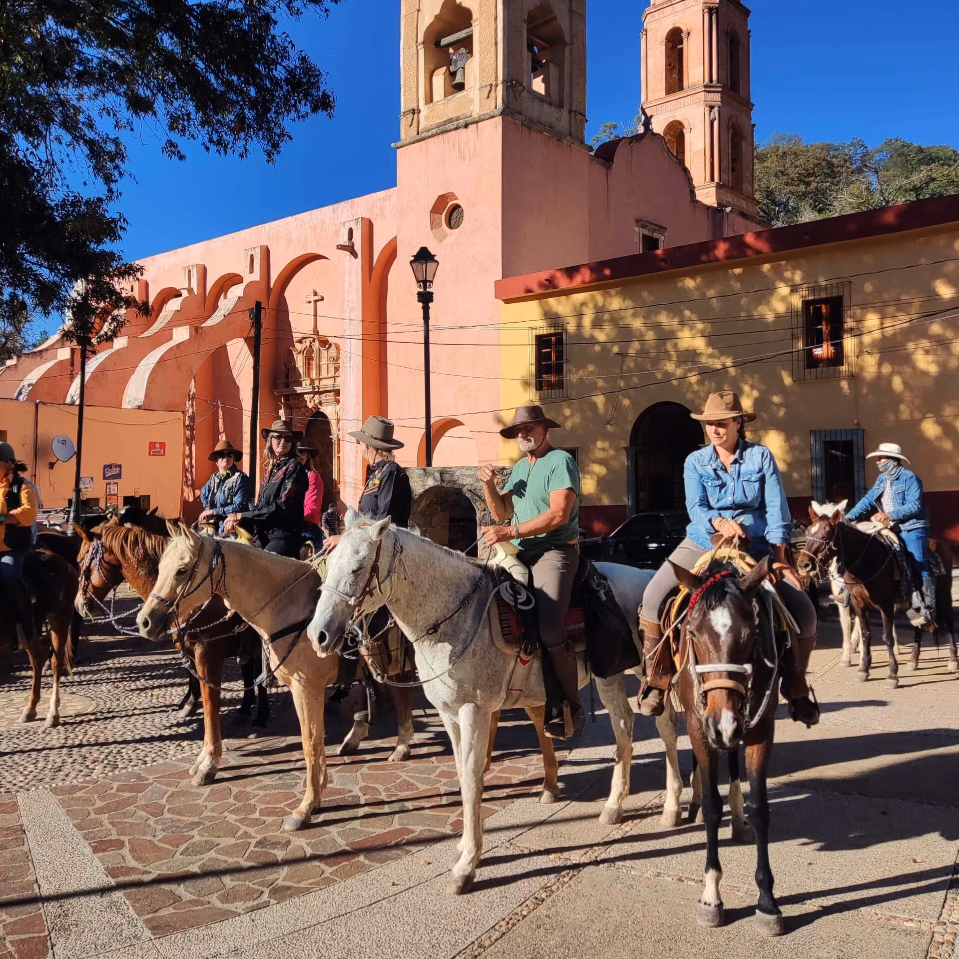Un groupe de cavaliers profite du soleil sur une place colorée d’un village mexicain, devant une église traditionnelle.