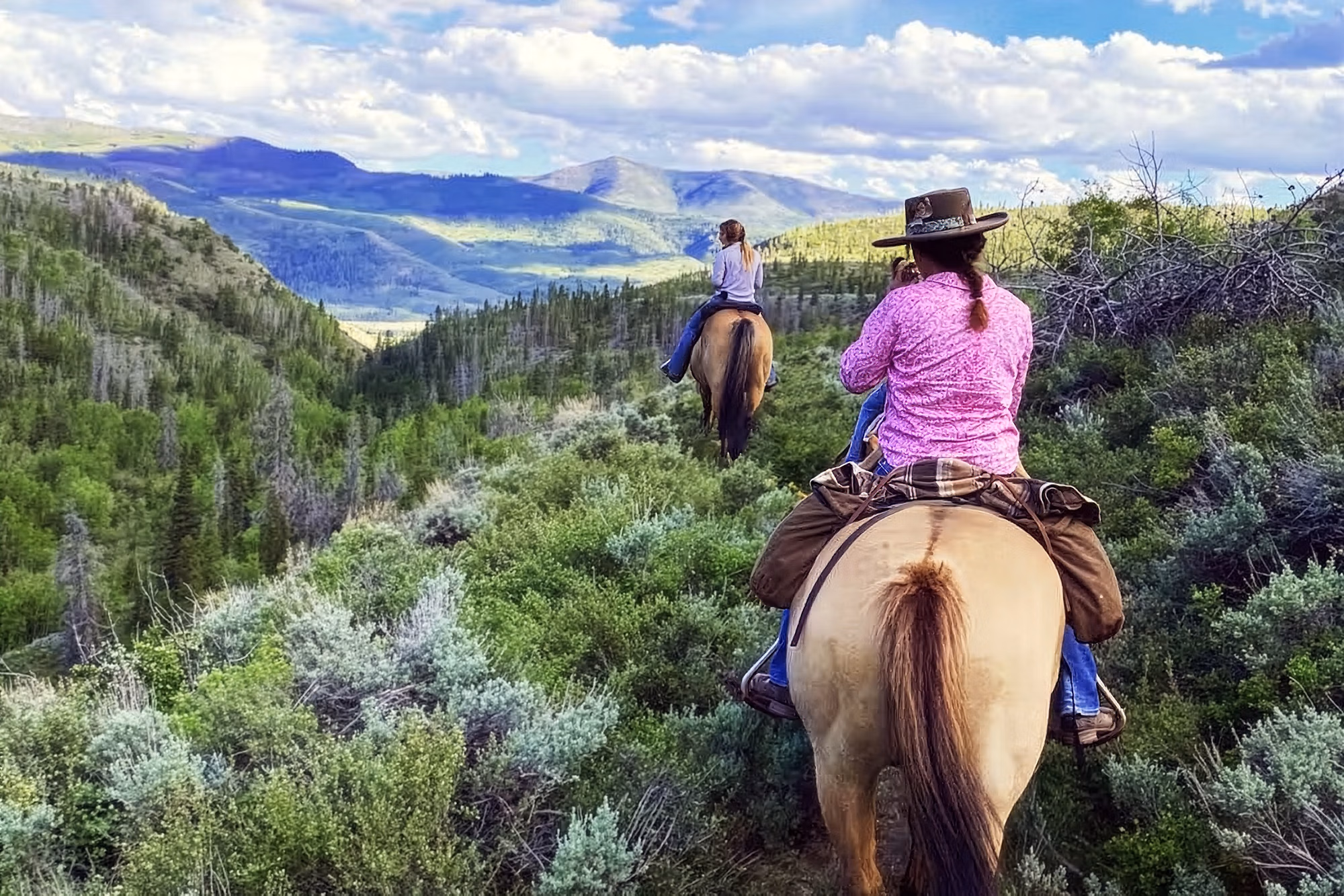 Deux femmes à cheval en pleine nature lors d’un séjour dans un ranch western, avec des forêts et des montagnes en arrière-plan.