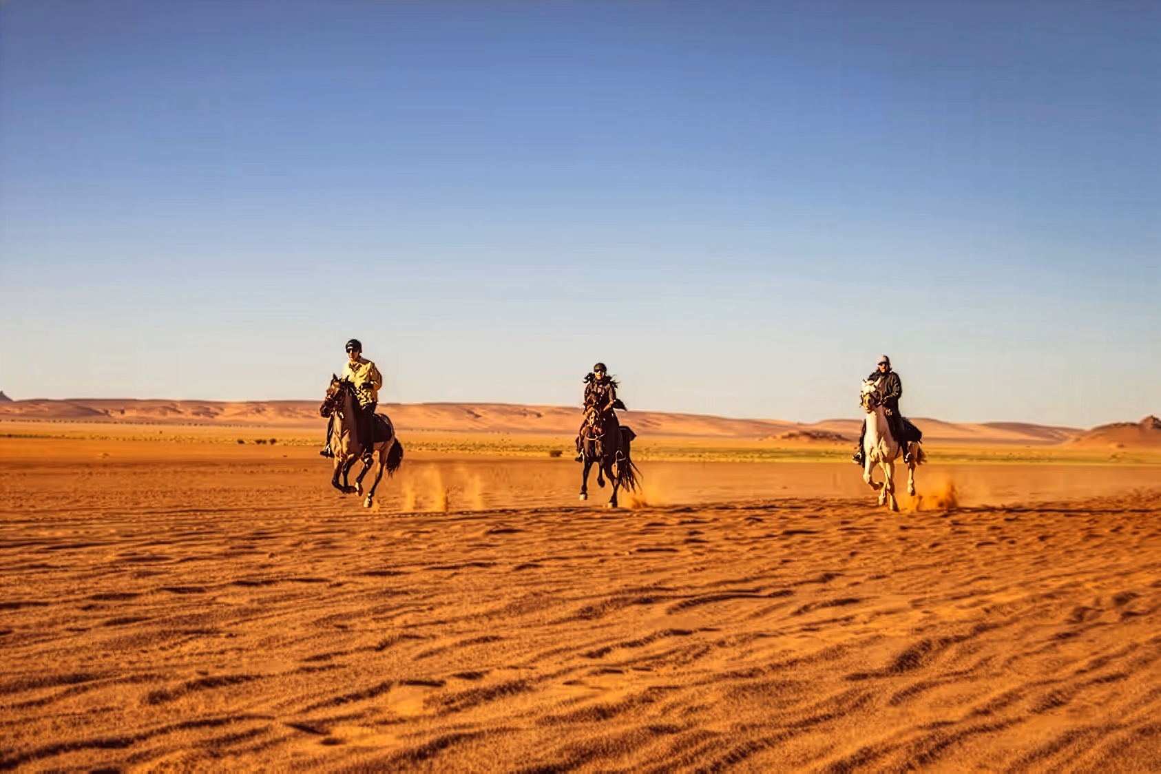 Trois cavaliers galopent à vive allure dans le Sahara au coucher du soleil.