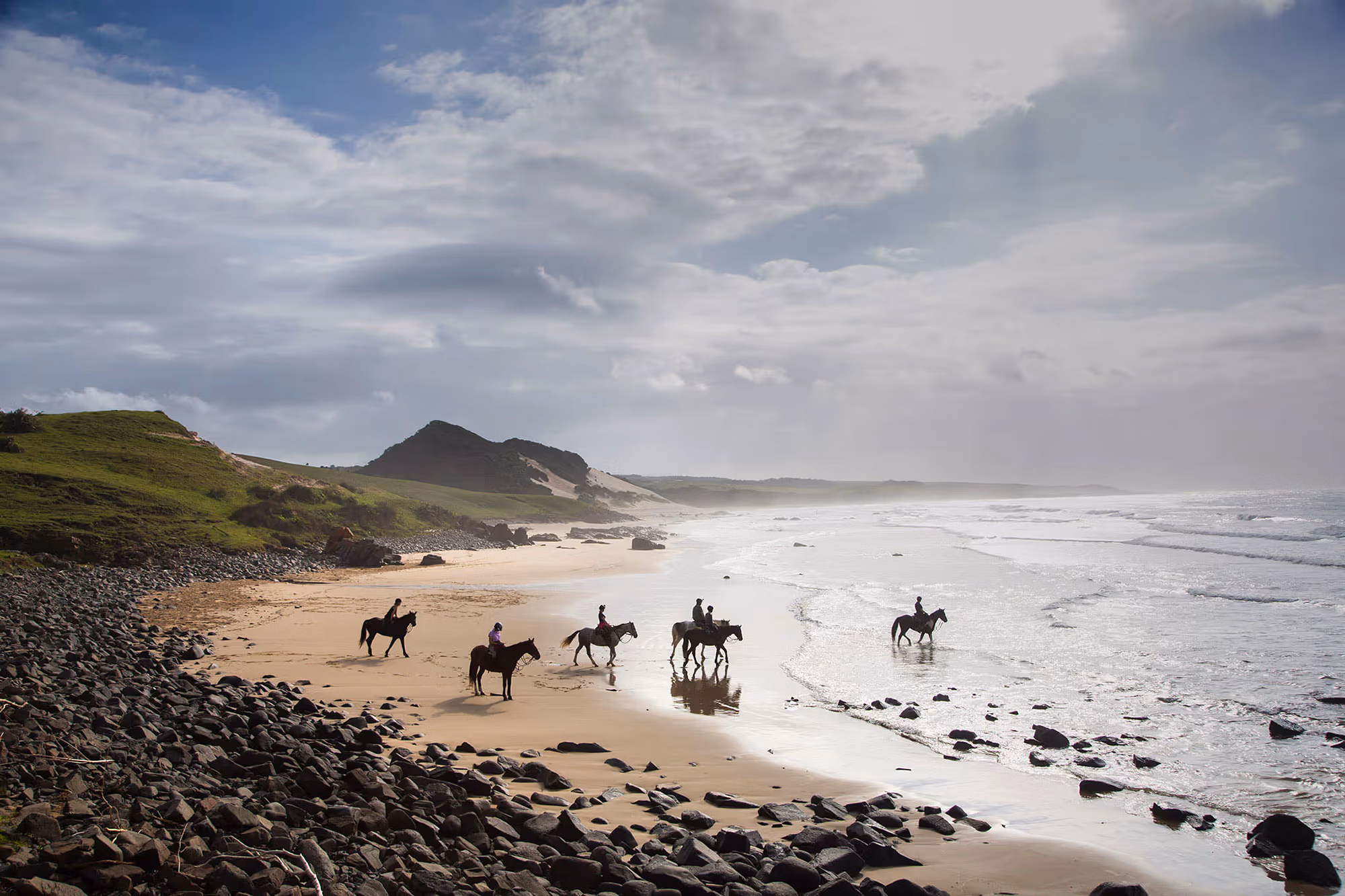 Un groupe de cavaliers explore une plage sauvage de sable et de rochers.