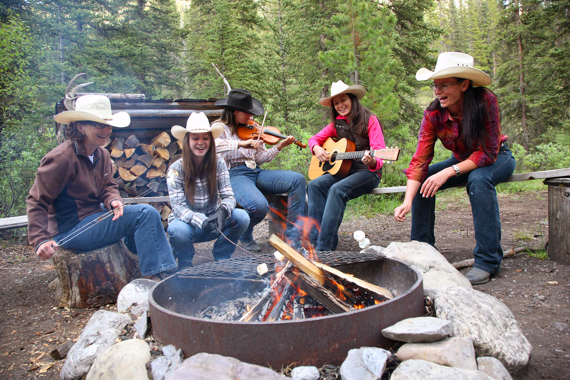 Un groupe de cavalières réuni autour d’un feu de camp en pleine nature, jouant du violon et de la guitare acoustique pendant une pause.