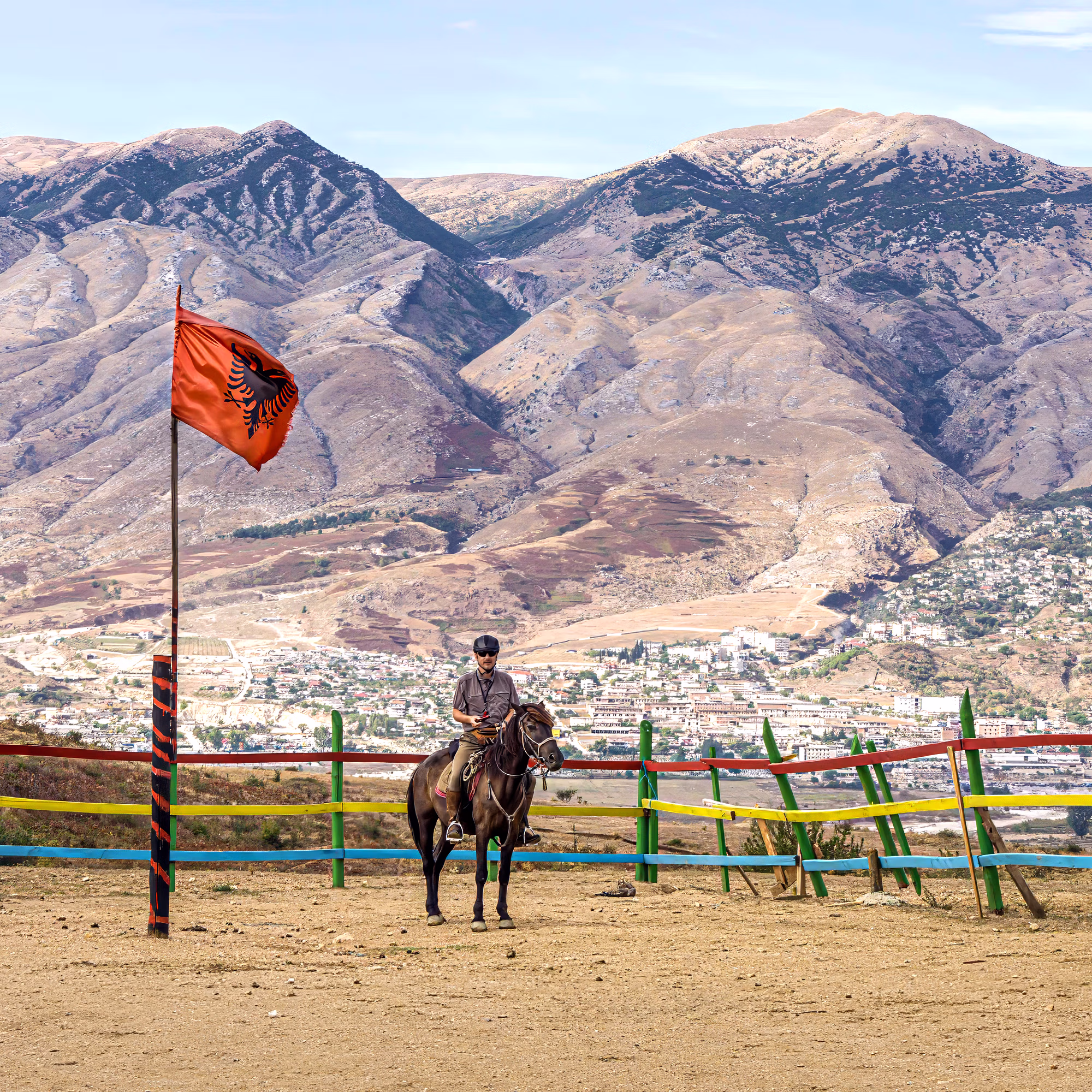 Un cavalier est assis sur son cheval dans un ranch traditionnel, avec une ville albanaise au loin en arrière-plan et un grand drapeau national flottant dans le vent.