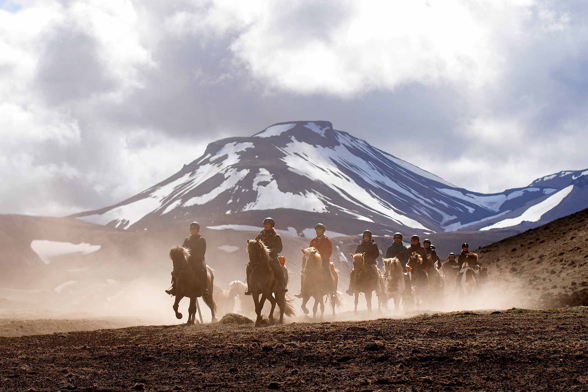 Cavaliers sur des chevaux islandais avançant sur un chemin accidenté devant une volcan enneigé lors d’un voyage à cheval en Islande.
