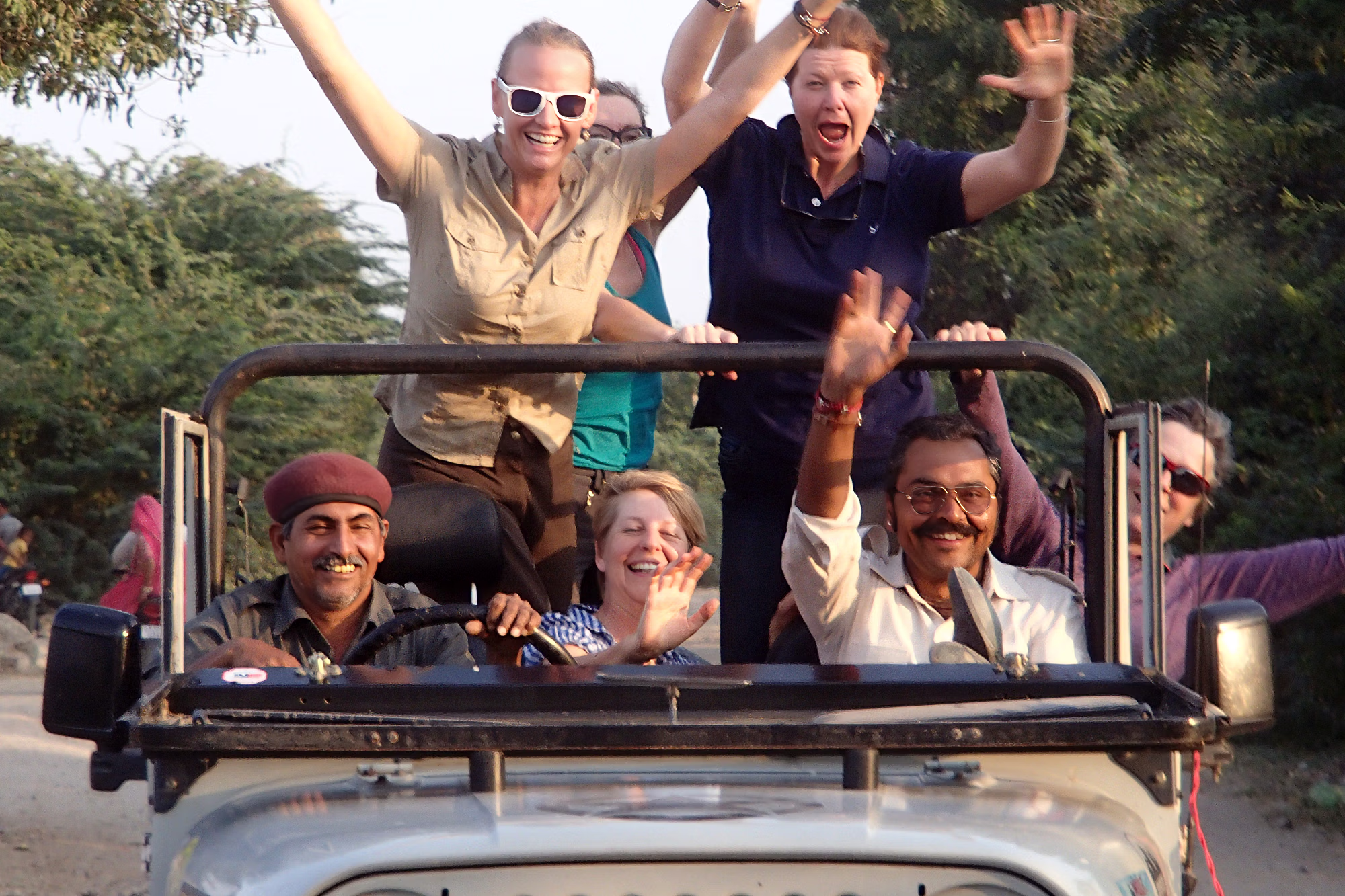 Des voyageurs et leurs guides locaux rient et lèvent les bras à bord d’une jeep de safari ouverte.