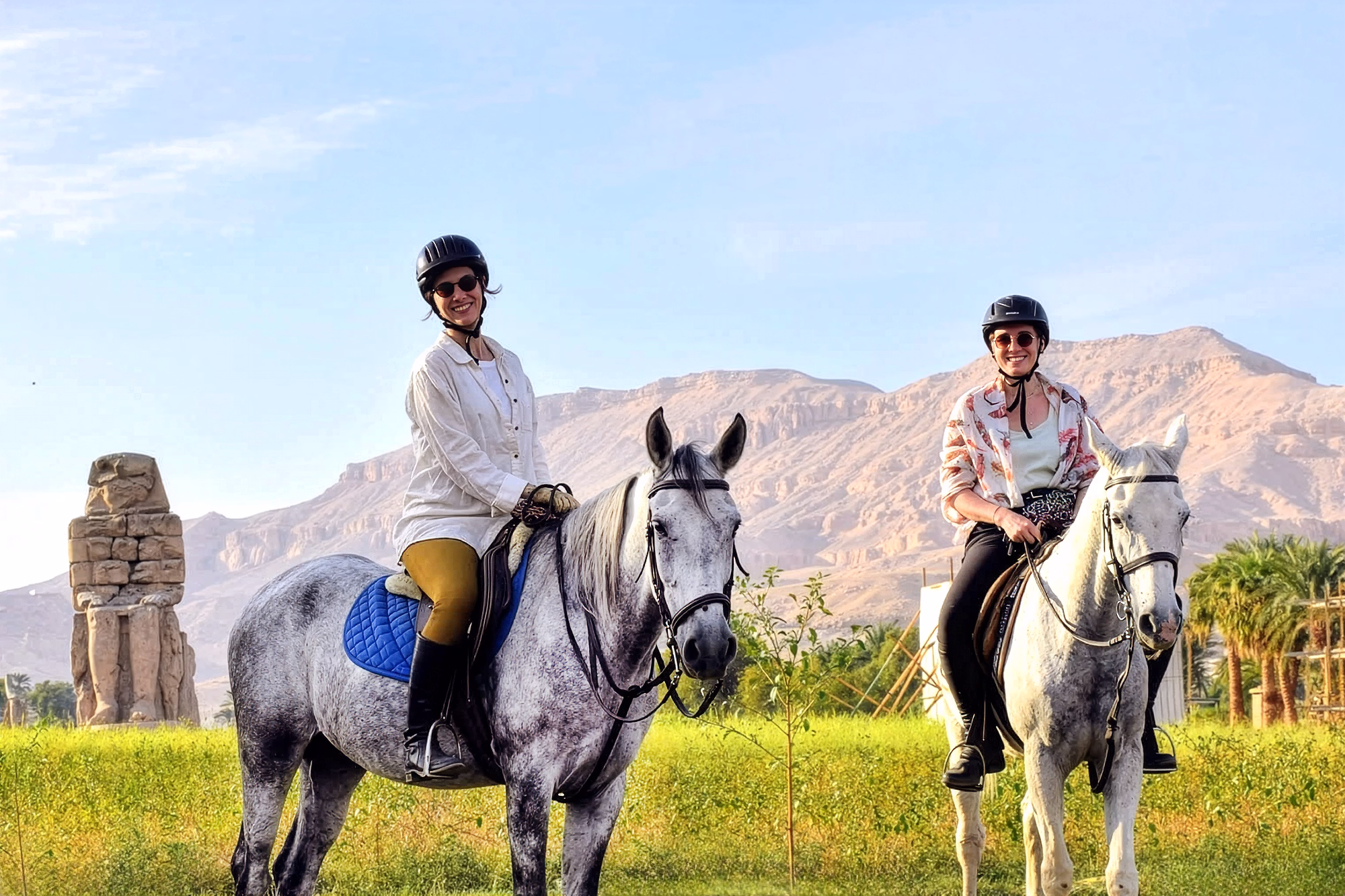 Deux cavalières souriantes posent à cheval dans la campagne verdoyante de Louxor, avec les montagnes en arrière-plan.