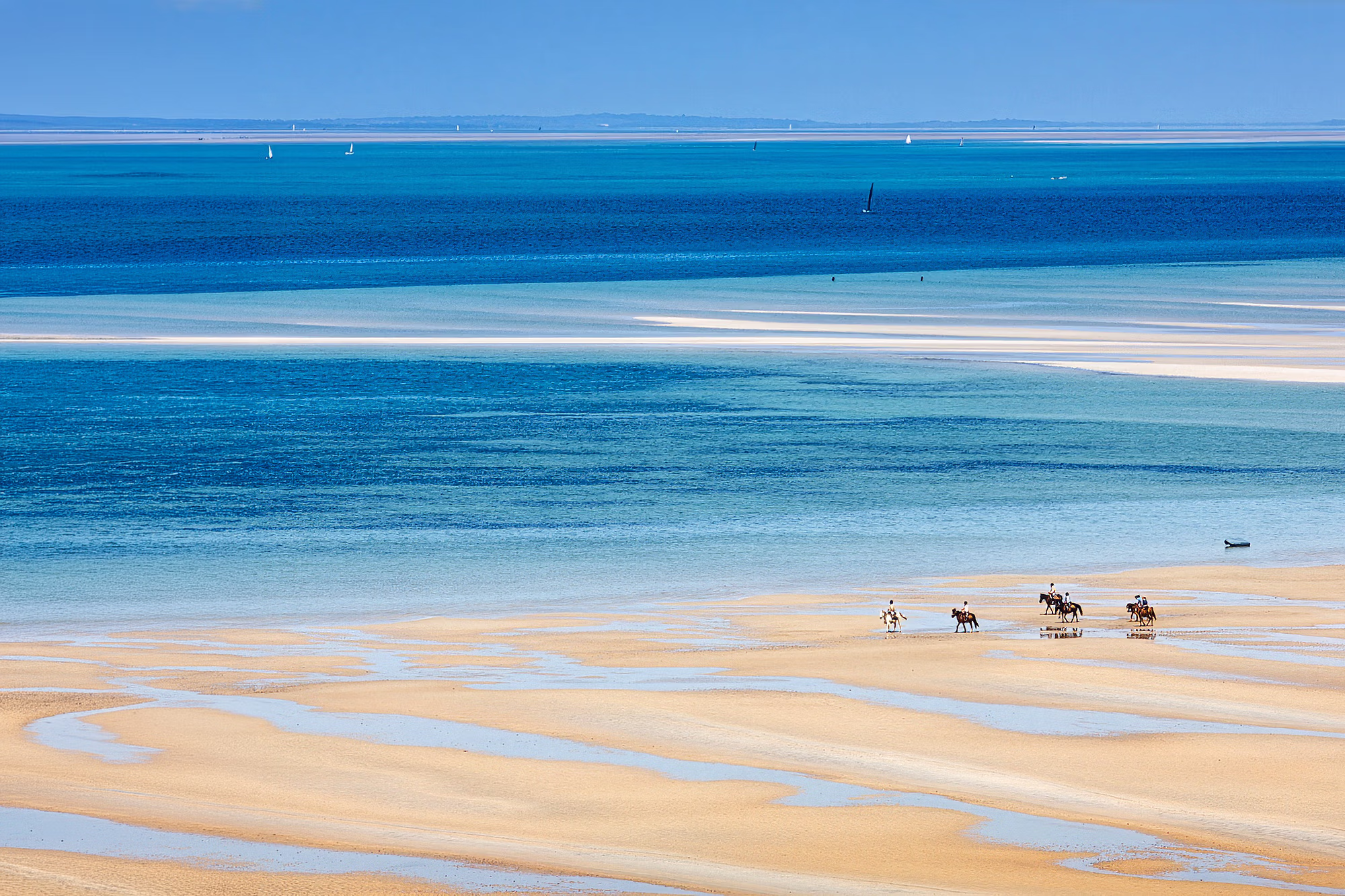 Un petit groupe de cavaliers traverse une vaste plage de sable blanc bordée d’eaux turquoise et de dunes.