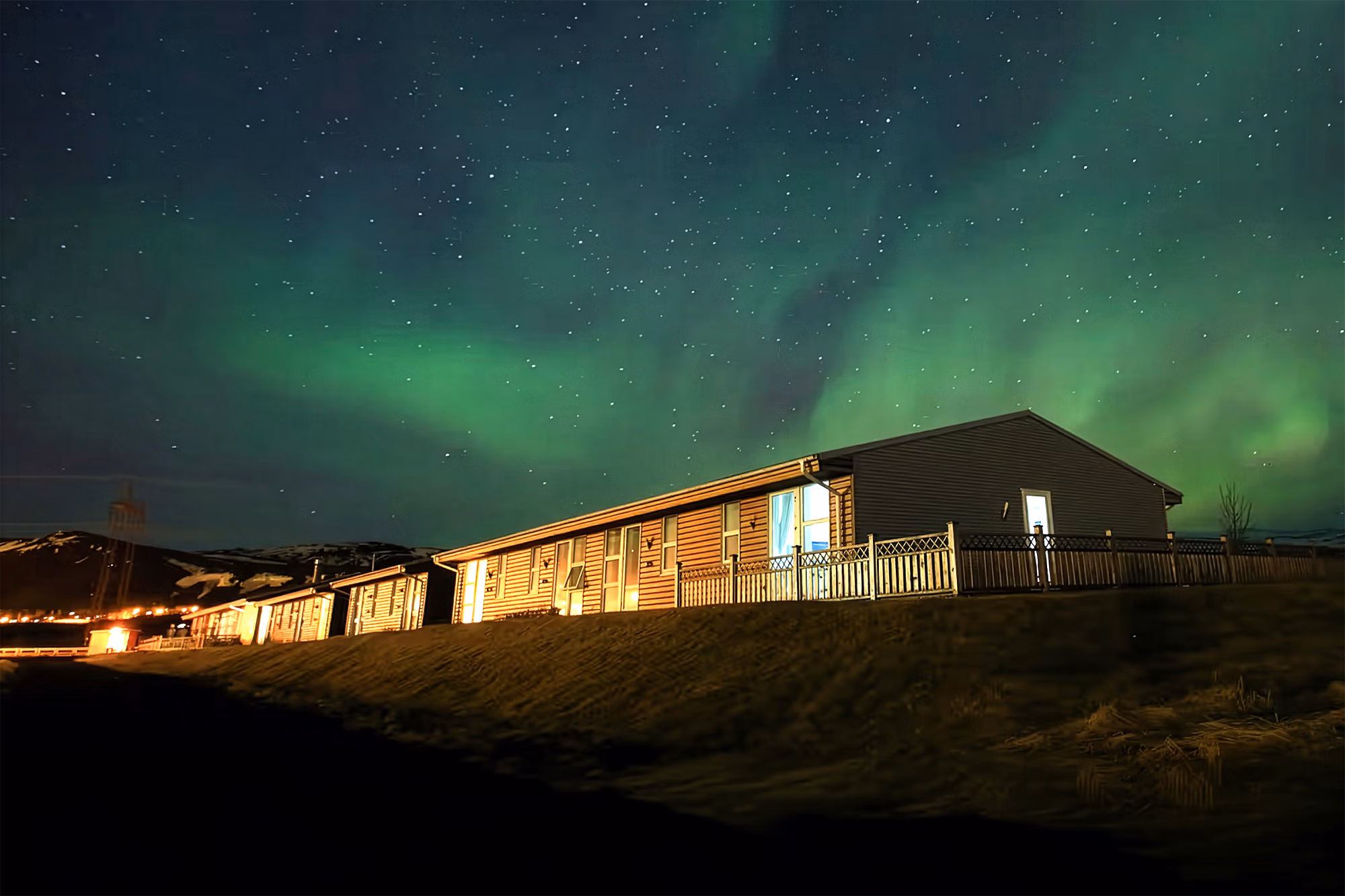 Une rangée de chalets chaleureux en hiver, aux fenêtres illuminées, sous un ciel arctique nocturne parcouru de magnifiques aurores boréales.