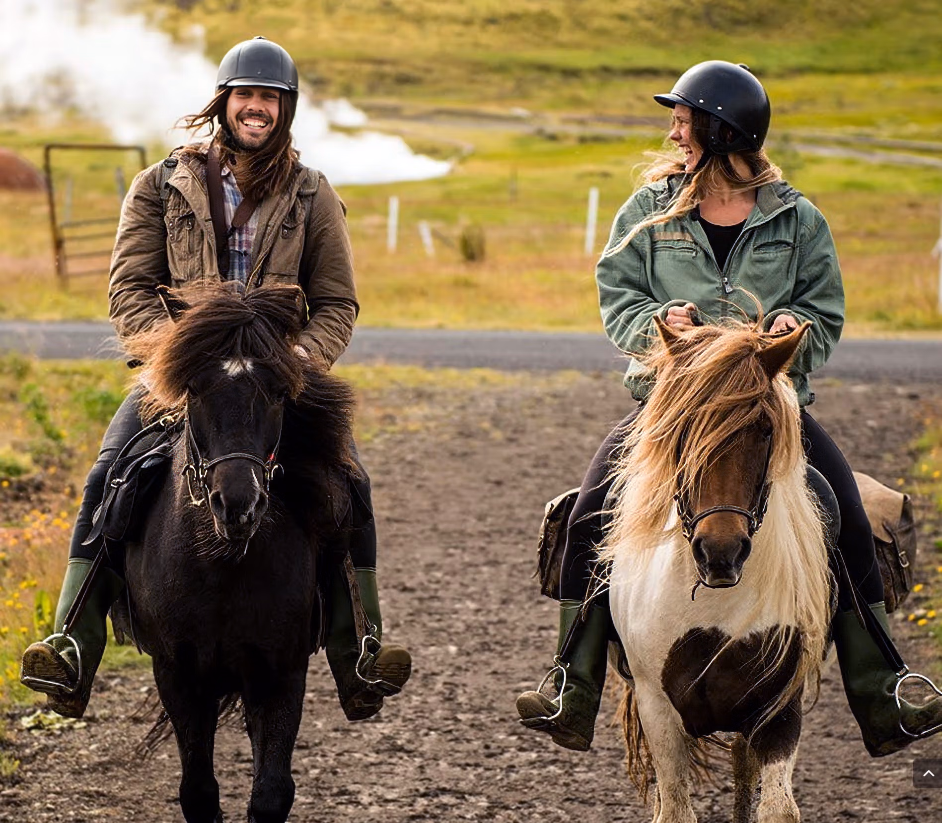 Deux cavaliers souriant et discutant à cheval devant une prairie verdoyante lors d’une randonnée équestre en Islande.
