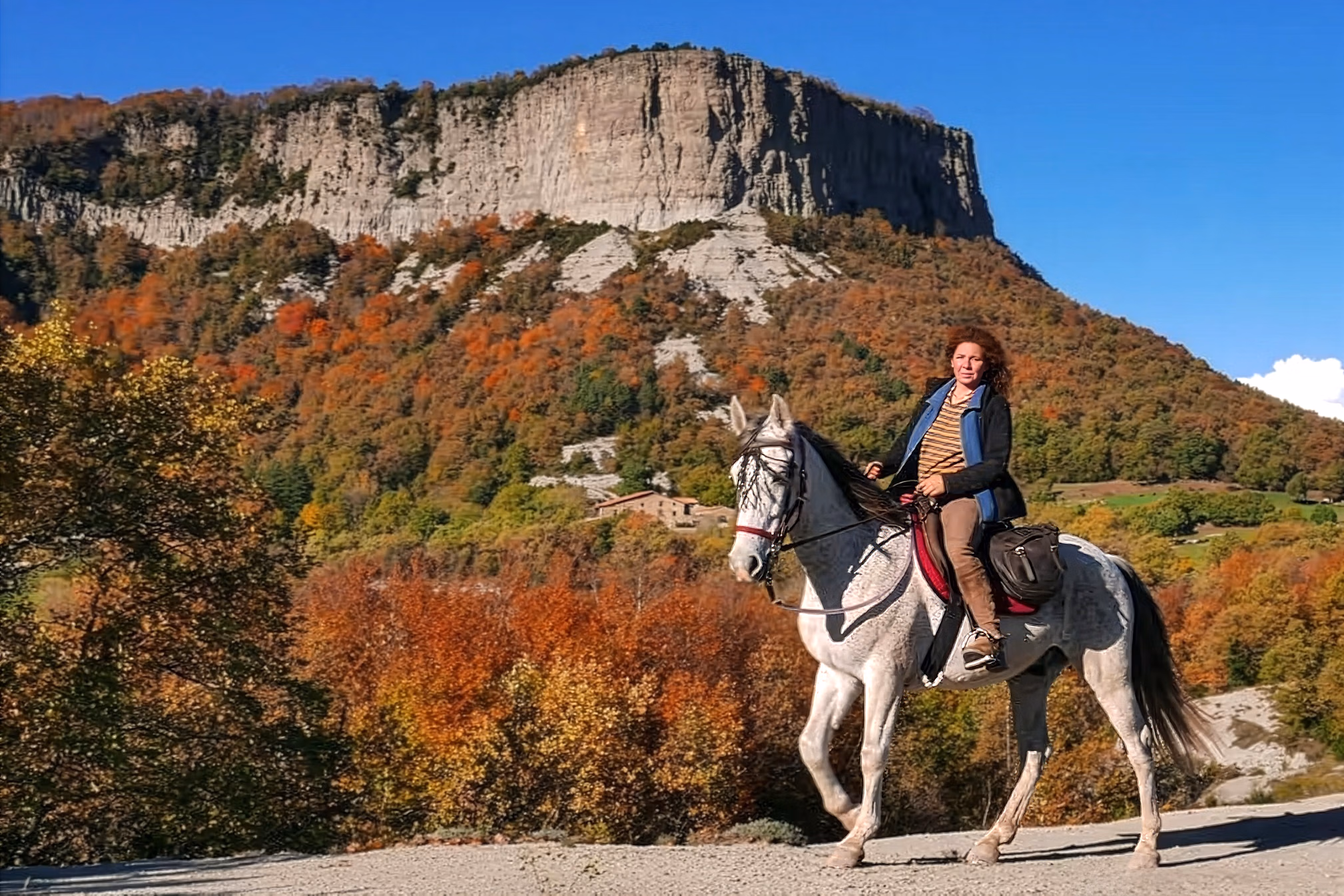 Une cavalière regarde vers l’objectif, avec une crête montagneuse et une forêt aux couleurs d’automne en arrière-plan.
