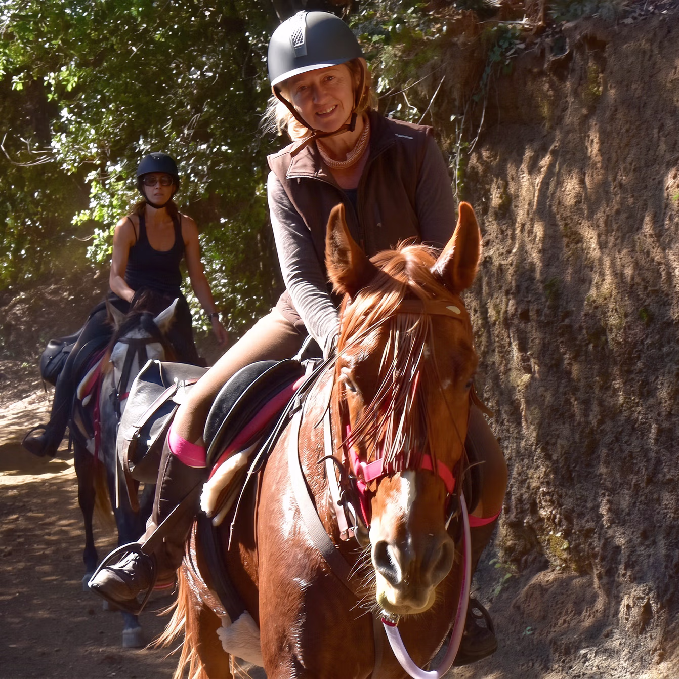 Deux cavalières avancent en file indienne sur un sentier, la cavalière souriante en tête.