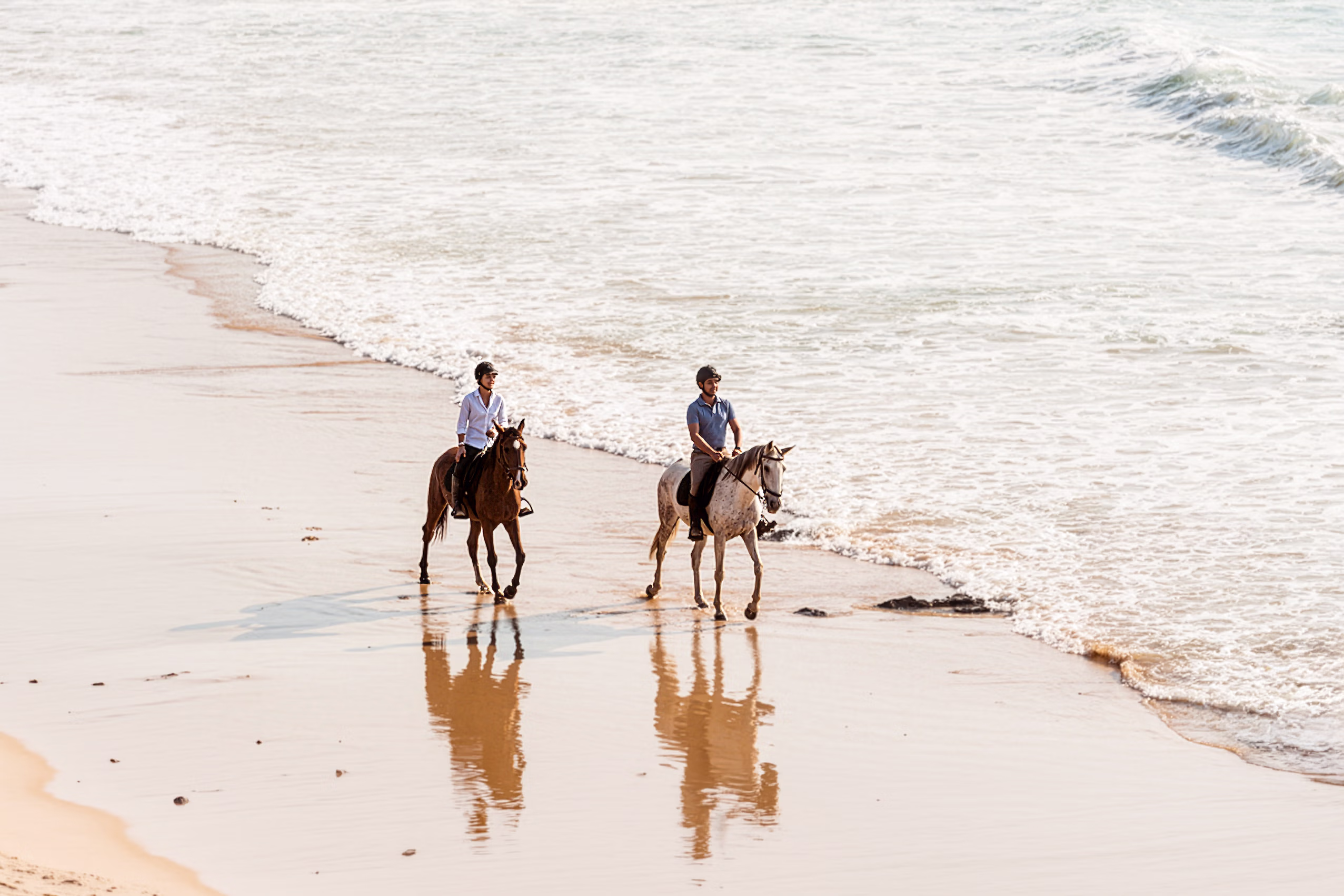 Deux cavaliers portant des casques profitent d’une balade à cheval sur une plage de sable de la côte de l’Alentejo, au bord des vagues.