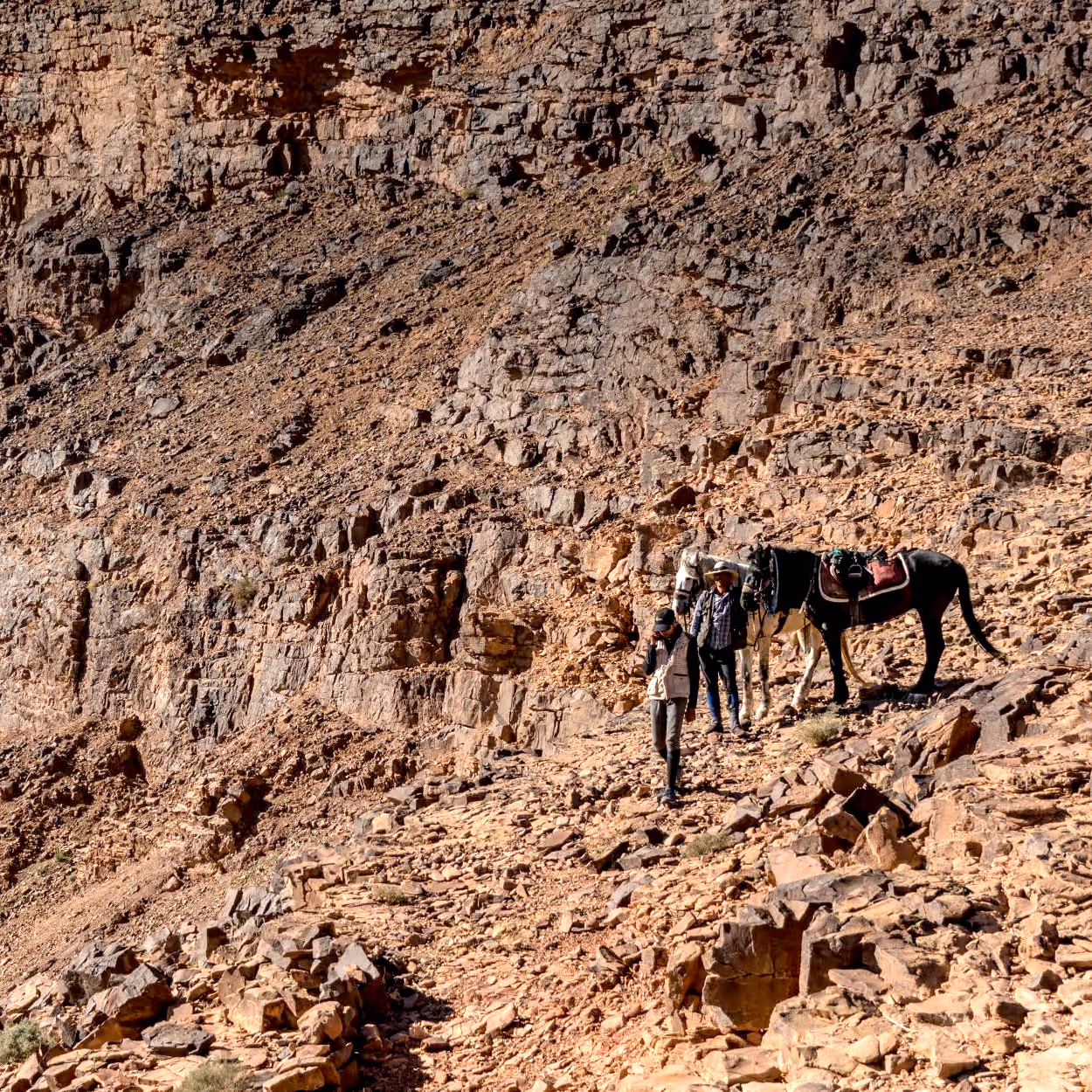 Un cheval et deux guides font une pause sur une pente rocheuse rouge et escarpée.