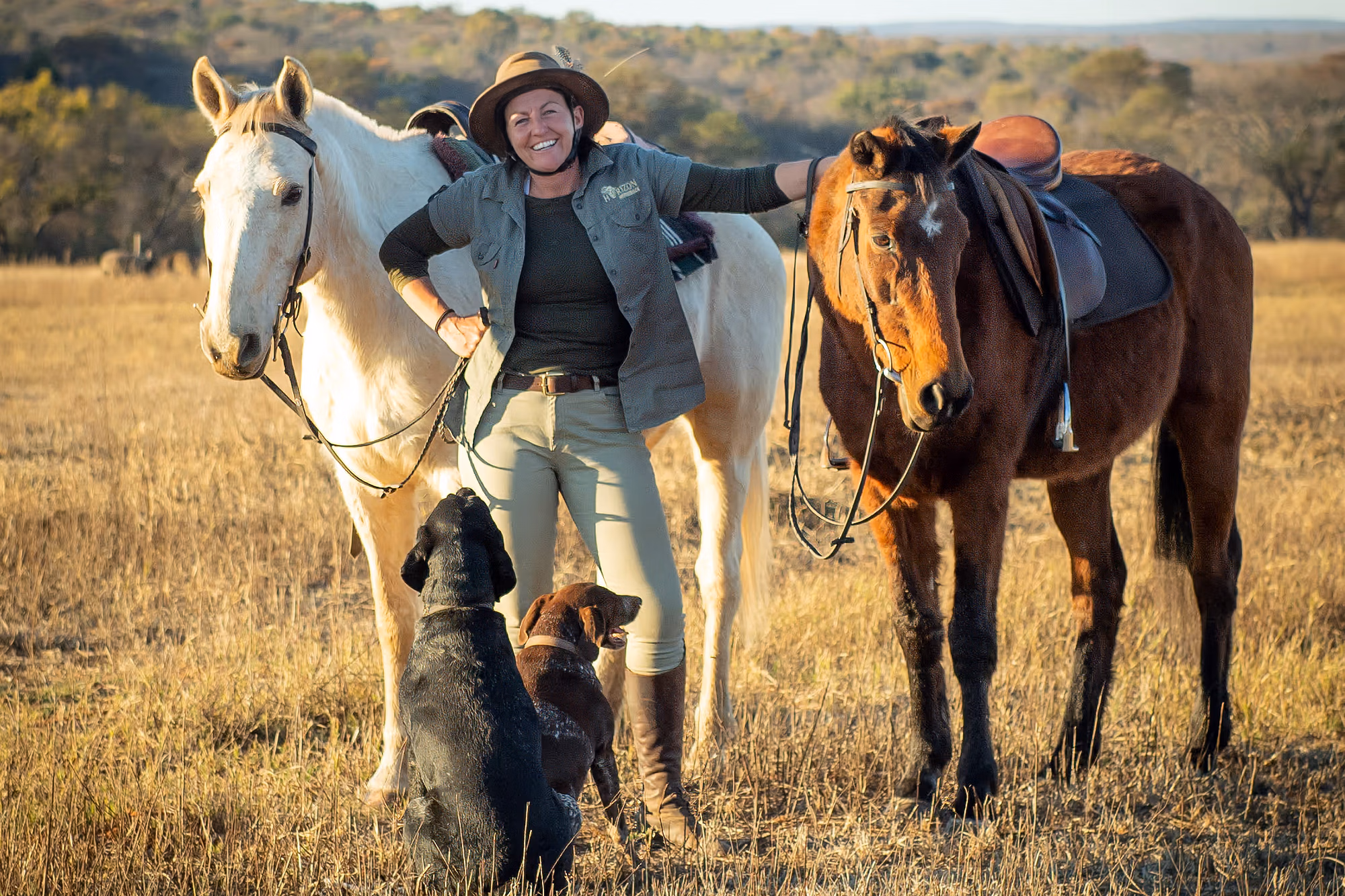 Cavalière debout devant deux chevaux avec son chien dans la brousse sud-africaine.