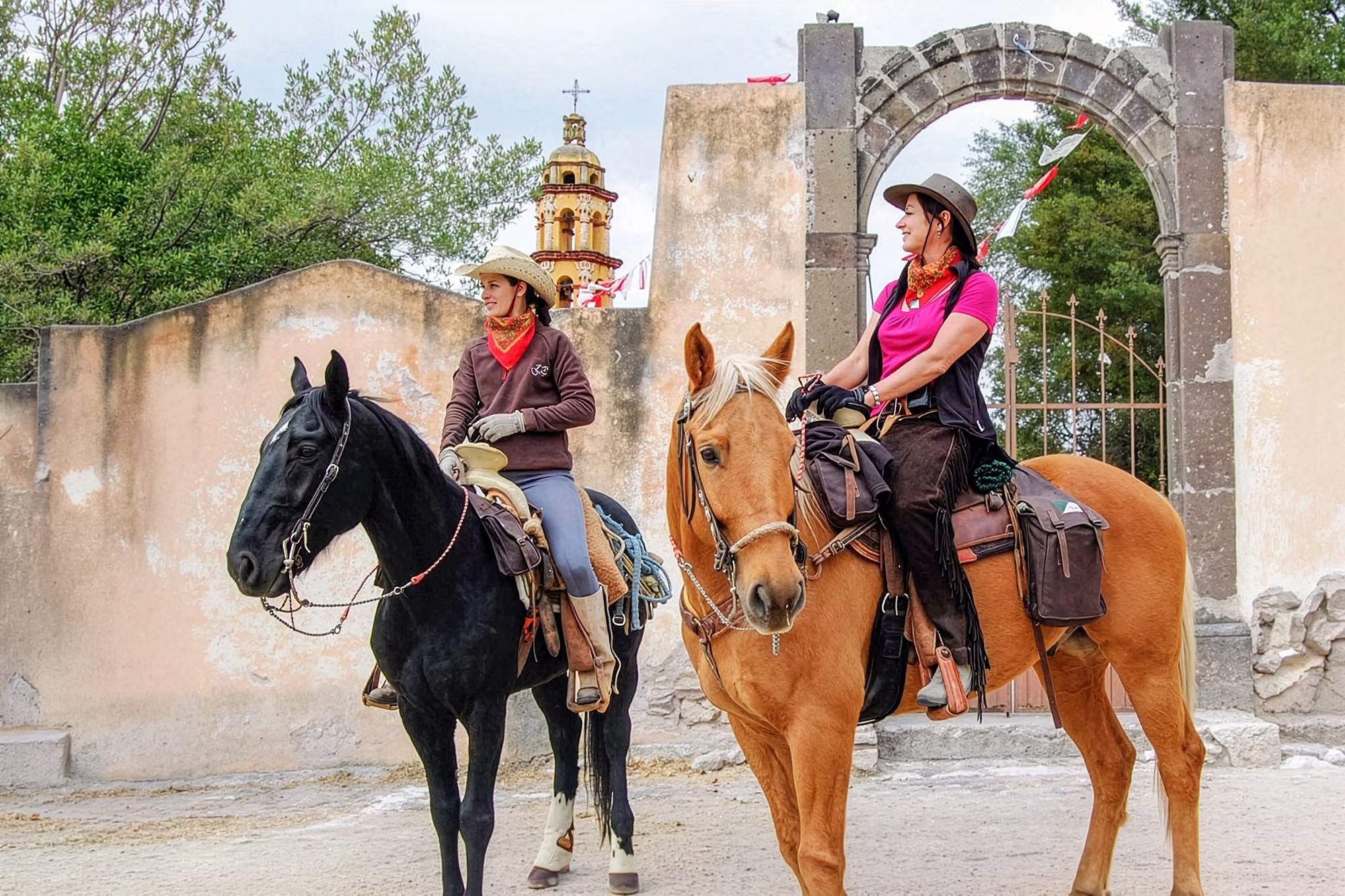 Des cavalières souriantes lors d’une randonnée équestre dans une ville traditionnelle mexicaine.