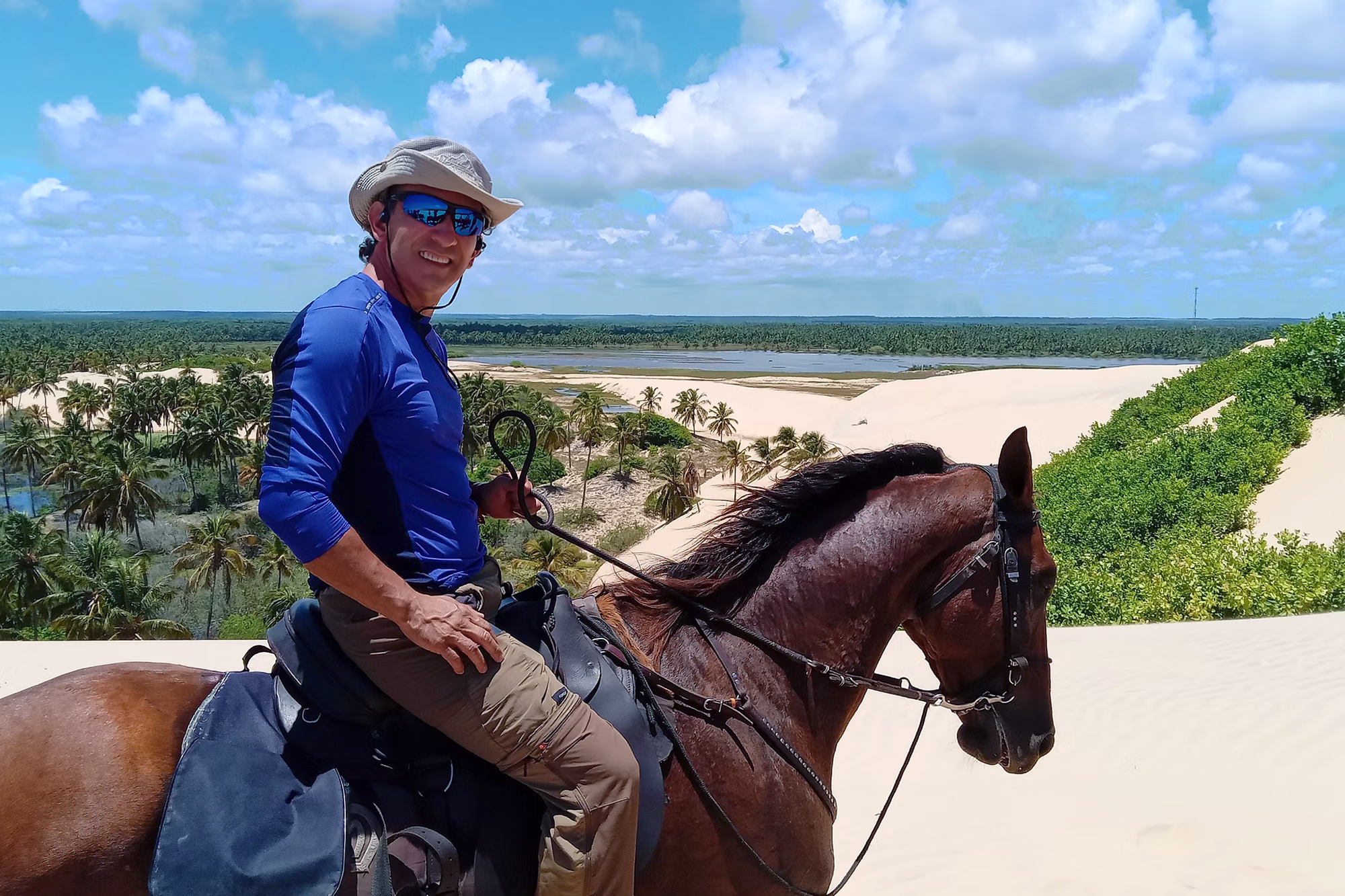 Un cavalier souriant regarde l’objectif en traversant une grande dune de sable à cheval.