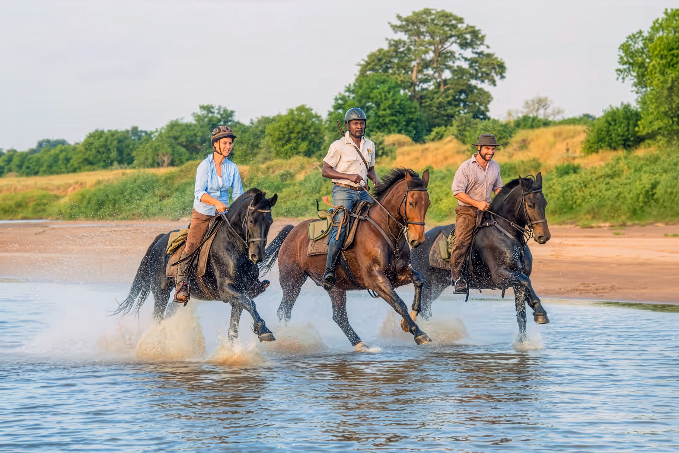 Trois cavaliers galopent dans l’eau en suivant le courant de la rivière.