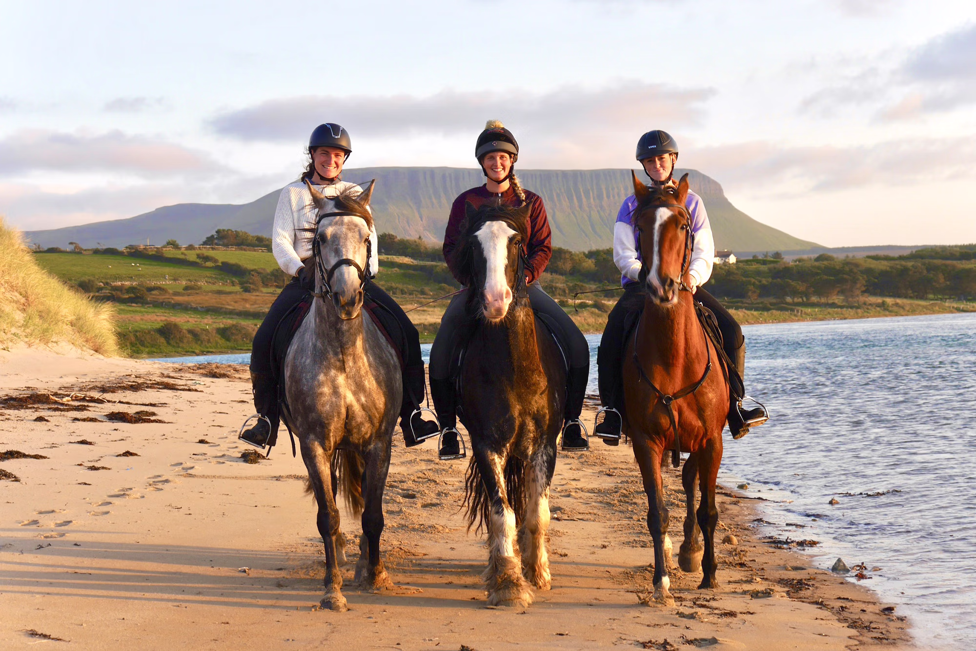 Trois cavaliers parcourent une vaste plage de sable, avec une imposante montagne en arrière-plan.