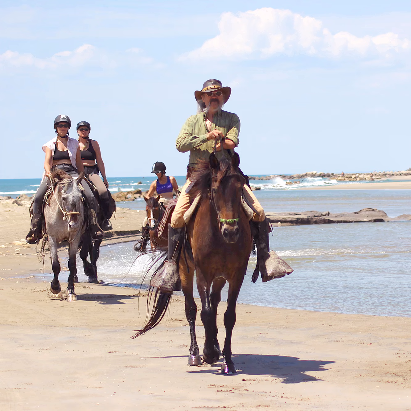 A small group of trail riders trotting their horses along the wide sandy coastline