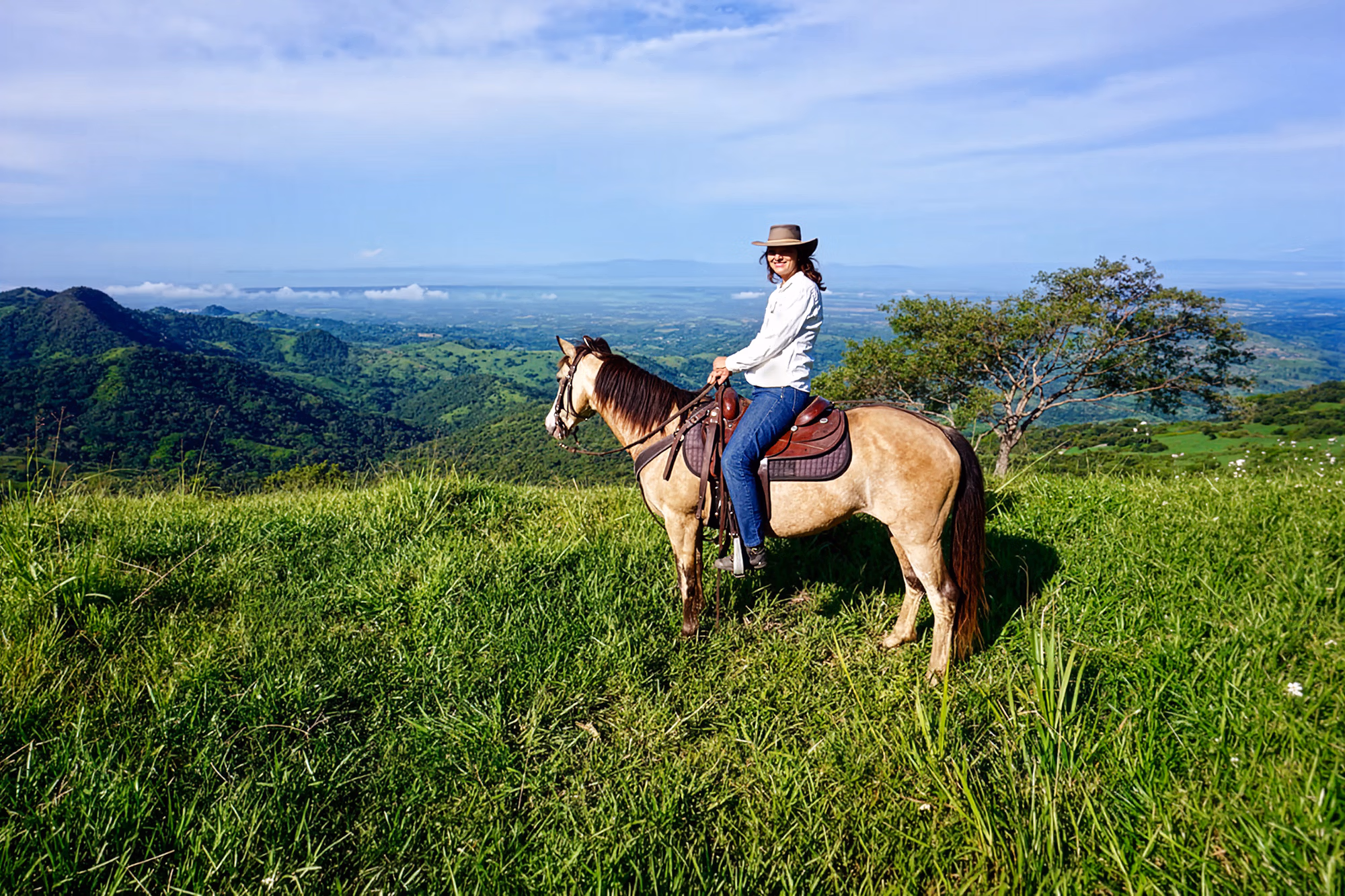 Une cavalière souriante portant un chapeau de cowboy s’arrête sur une colline verdoyante lors d’une randonnée sur le Golden Hills Trail au Costa Rica.