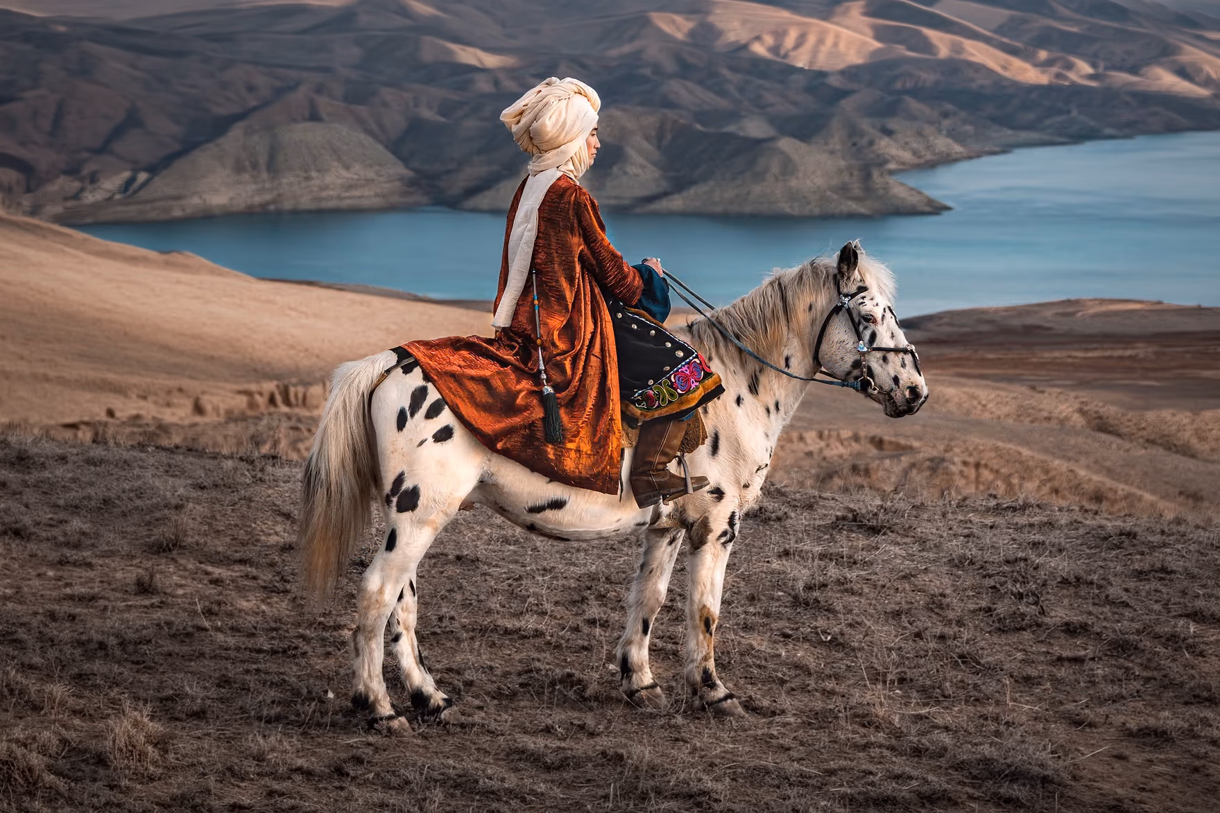 Une femme en tenue traditionnelle monte un rare cheval Chaar Appaloosa et contemple les paysages uniques du Kirghizistan lors d’une randonnée nomade.