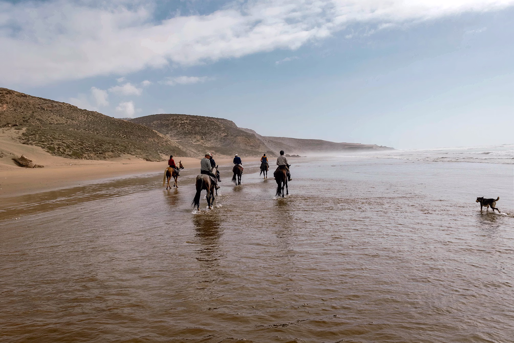 Un groupe de cavaliers et un chien avancent dans les eaux de l’océan Atlantique devant de belles dunes côtières.