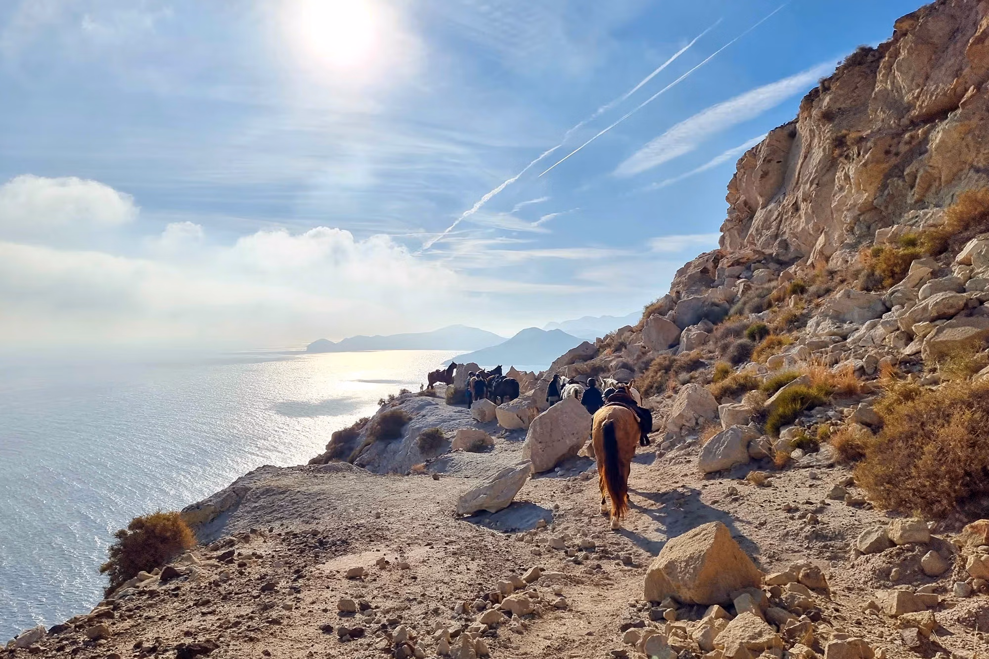 Une file de cavaliers suit un sentier rocheux bordé d’imposants rochers, avec vue sur la mer.