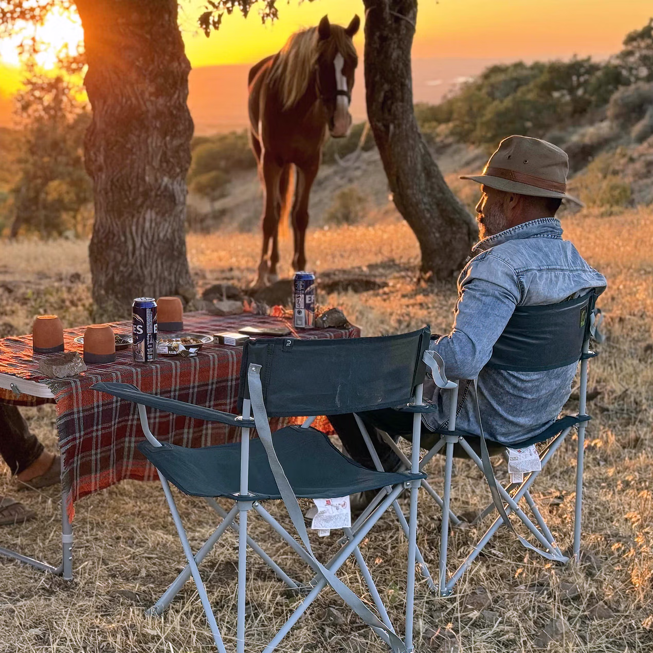 Deux cavaliers sont assis sur des chaises pliantes au coucher du soleil, avec un cheval derrière eux sous un ciel doré et rouge.