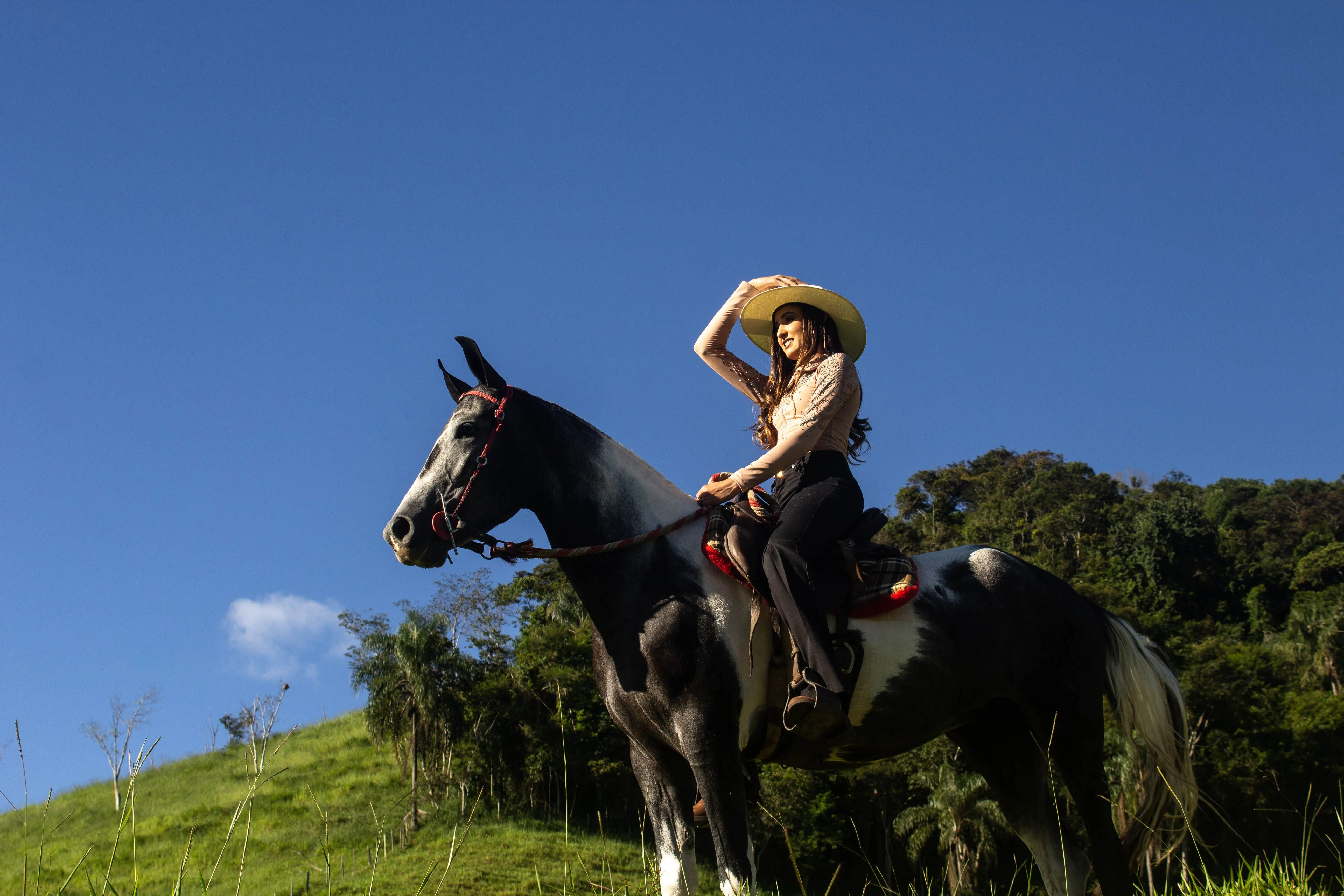 Une cavalière souriante sur un cheval pie dans un paysage verdoyant sous un ciel bleu.